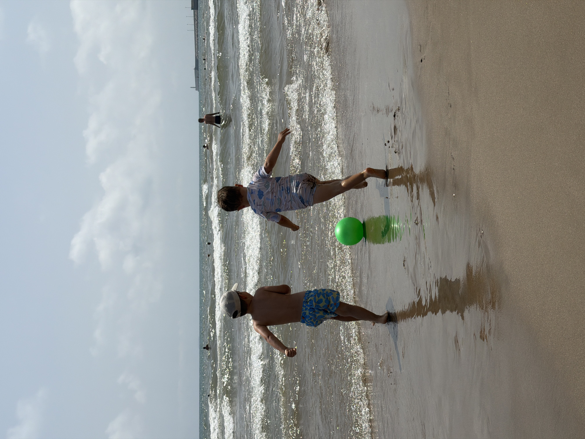 Two young boys are playing on a sandy beach with a green ball near the shoreline. The boy on the left, wearing a cap and patterned swim trunks, faces away from the camera. The boy on the right, in a light-colored swimsuit with a pattern, also faces away. The sea has moderate waves and other people are visible swimming and wading in the water. In the distance, a breakwater extends into the sea, with a lighthouse at its end. The sky is partly cloudy.