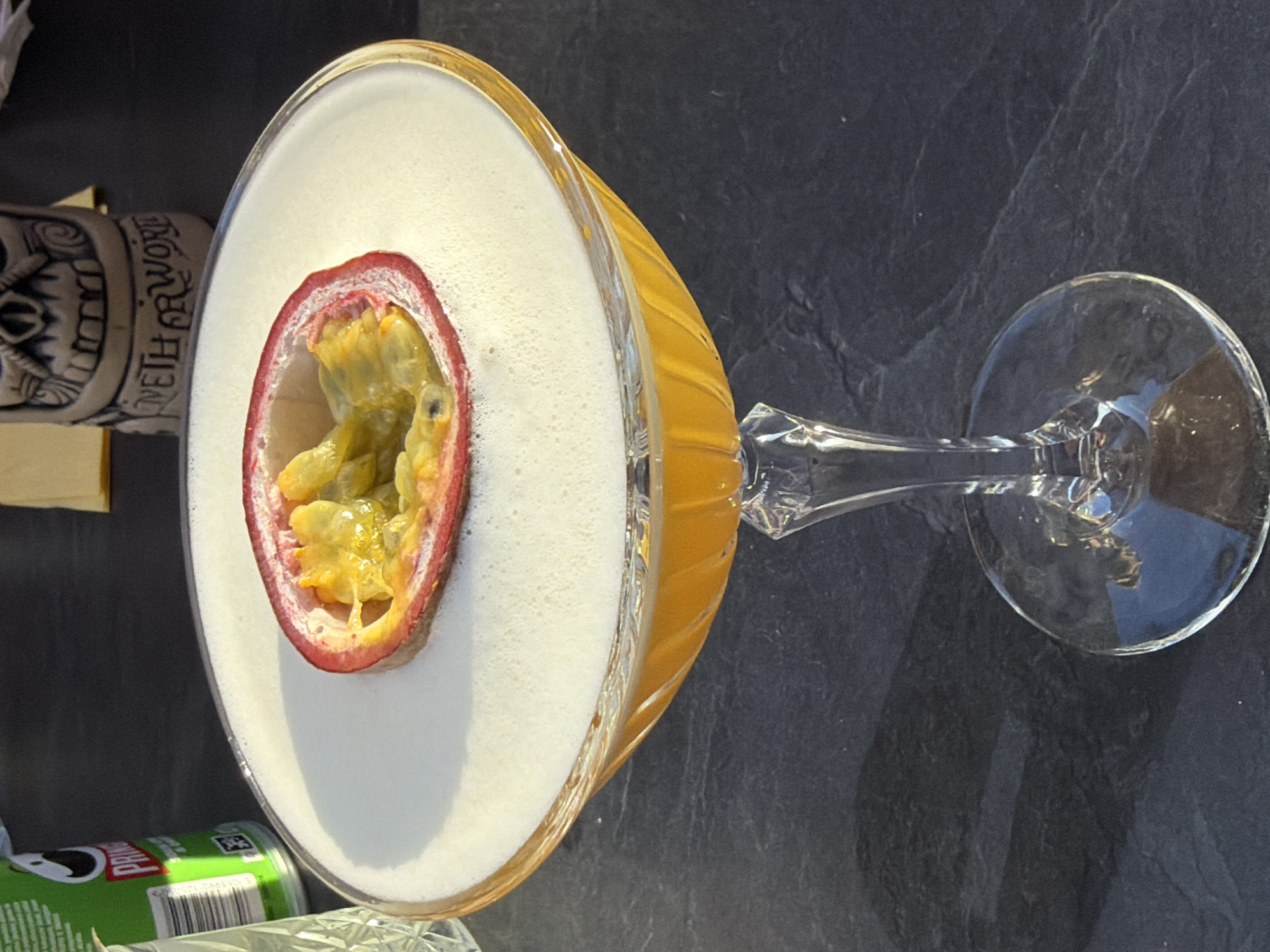 An overhead shot shows a cocktail in a clear glass coupe on a dark gray slate surface. The cocktail appears to be topped with a thick, foamy white layer, garnished with half a passion fruit, revealing the yellow pulp and black seeds. The cocktail itself has an amber hue. A tiki-themed ceramic mug and a can of Pringles chips are partially visible in the lower left corner of the image. The setting resembles a table or countertop, likely within a bar or restaurant. There is no indication that the location is in Riga, Latvia.