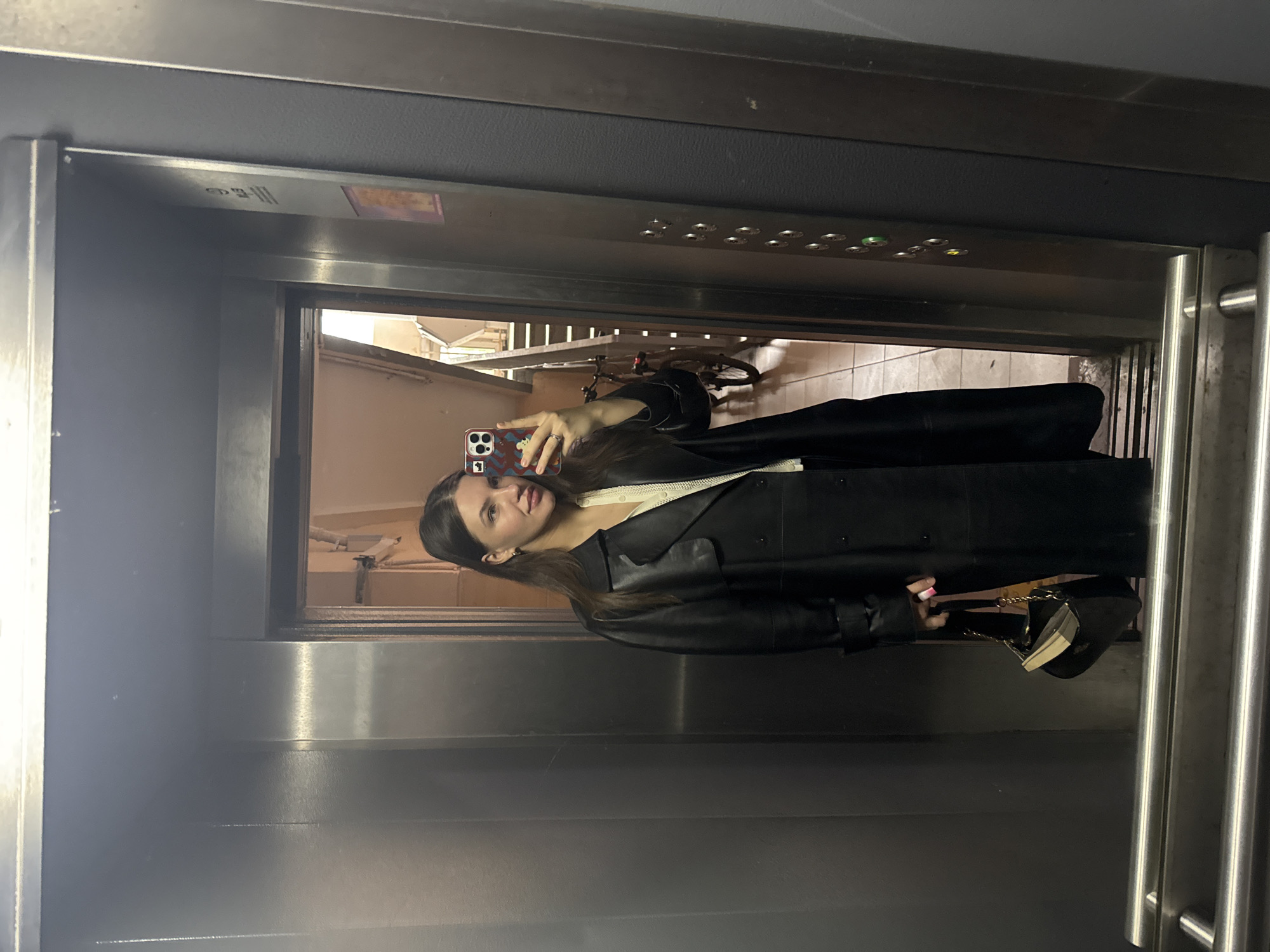 A woman is taking a selfie in a metal elevator. The elevator is a mirror. The elevator door is open. A bicycle and stairs are visible behind the woman. The woman is wearing a long black coat and carrying a small black purse with a gold chain. The location is likely within Moscow, Russia.
