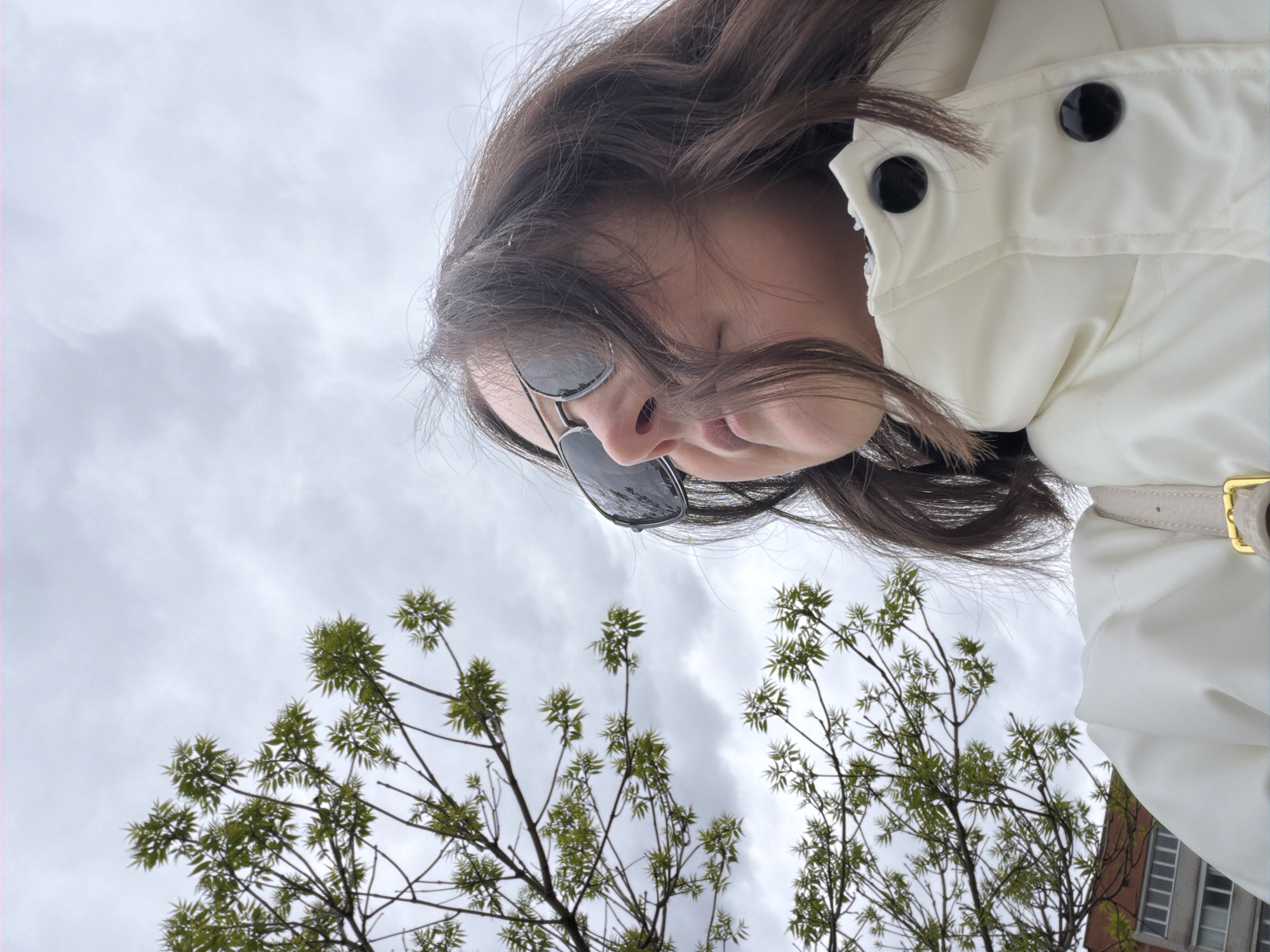 A low-angle shot shows a woman with dark hair and sunglasses looking down. She is wearing a white jacket with black buttons. The background consists of tree branches with new green leaves against a cloudy sky. A building with windows is partially visible in the lower right corner. The location is an urban area, possibly a city street.