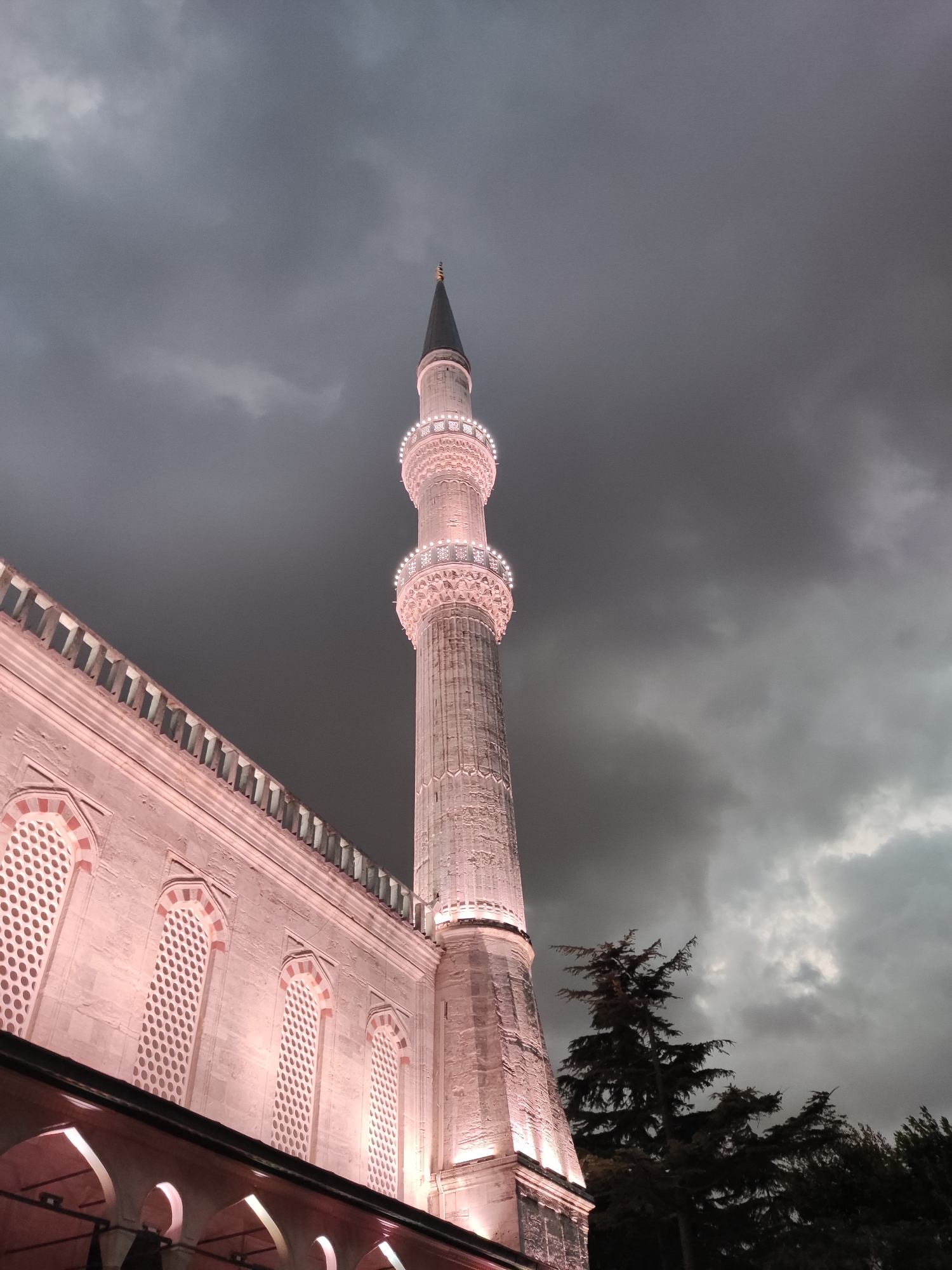 A low-angle, full shot captures the exterior of the Sultan Ahmed Mosque, also known as the Blue Mosque, in Istanbul, Turkey. The minaret, illuminated by pink lights, stands tall against a dark, cloudy sky. The stone facade of the mosque is also lit, highlighting arched windows with intricate lattice work. Trees are visible to the right of the minaret, their dark silhouettes contrasting with the illuminated architecture. The image is taken at dusk or night, indicated by the dramatic sky and artificial lighting.