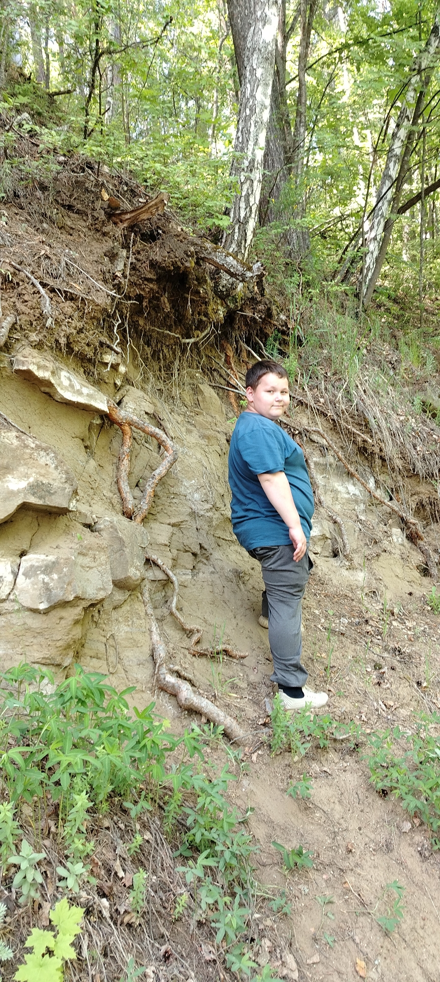 A young person in a teal t-shirt and grey pants stands in a natural outdoor environment. The person is positioned against a light brown, layered rock formation with exposed tree roots. Green vegetation is visible in the foreground and on the slope above the rock. The background consists of trees with green foliage. The location is near Noviy, Russia.