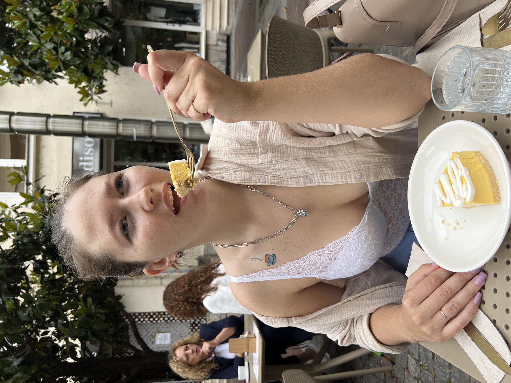 A fair-skinned woman with light brown hair eats dessert outdoors at a cafe. She holds a fork with a bite-sized piece of yellow cake towards her mouth. She wears a white lace bra, a silver chain necklace, and a light beige button-down shirt. The cafe table holds a plate with the remainder of her cake, a glass of water, and flatware. Two other women sit at the table behind her. The cafe sits on a sidewalk near a tree, and the storefront of a business called "diso" is in the background. The cafe is in the city of Podgorica, Montenegro.
