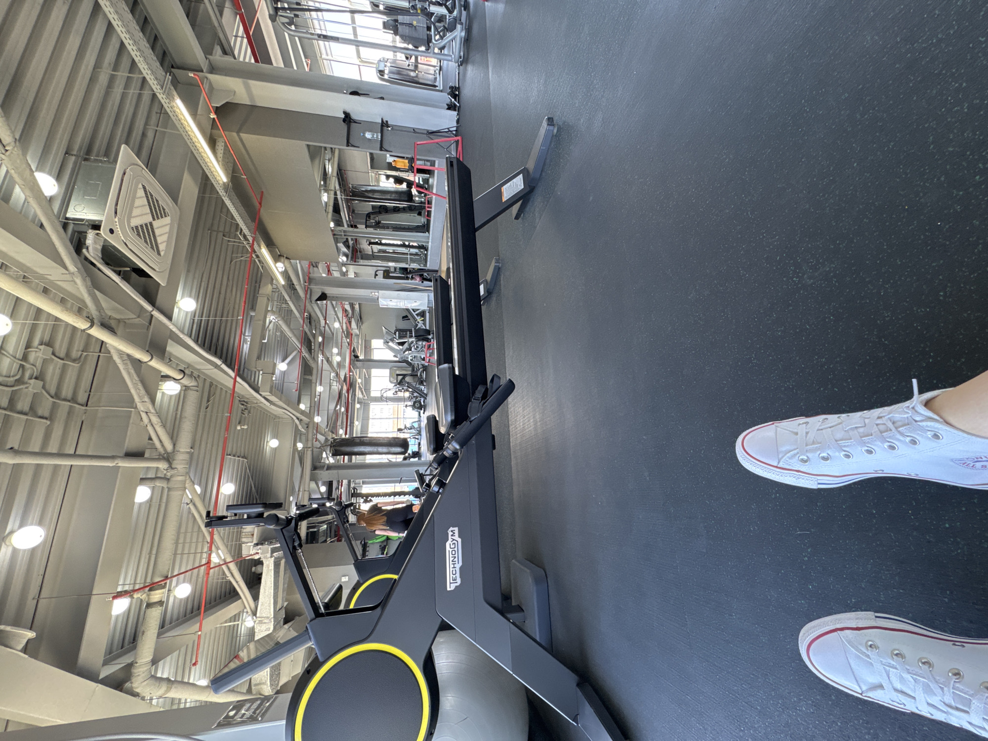 This media file presents a brightly lit, modern gym interior, captured with a 90-degree clockwise rotation. In the foreground, the feet of an individual wearing white Converse All Star sneakers are visible, standing on a dark grey, speckled rubber floor.
A prominent dark grey Technogym rowing machine, featuring circular yellow accents, dominates the midground. Beyond it, various other gym equipment, including weight machines and a red exercise step, are visible in the background, some reflected in large mirrors.
The gym's ceiling is high, showcasing an industrial design with exposed white pipes, ventilation ducts, and both linear and circular light fixtures. Red safety conduits or electrical cables traverse the ceiling. A white ceiling-mounted air conditioning unit with a geometric grille is also present.
The scene suggests an individual present in a fitness center, either beginning or concluding a workout. The gym appears well-maintained and features contemporary equipment. Visible text includes "TECHNOGYM" on the rowing machine and the "CONVERSE ALL STAR" logo on one of the sneakers. This gym is located in Belorechensk, Russia.