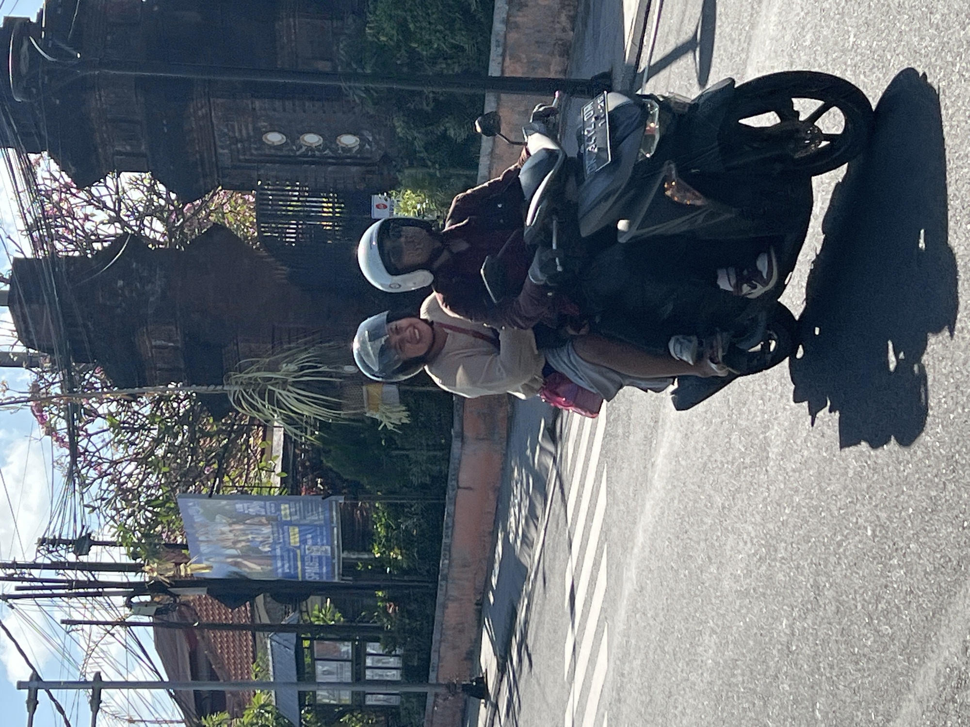 Two individuals, both wearing helmets, are riding a dark-colored scooter on an asphalt road. The person in front is driving, wearing a dark jacket and gloves. The person behind is a passenger, wearing a light-colored shirt and holding a pink bag. They are riding past a crosswalk. In the background, there is a brick wall topped with greenery and a large, ornate brick structure, possibly an entrance gate. Overhead, a dense network of electrical wires is visible against a blue sky with scattered clouds. Signs and trees are also present in the background, indicating an urban or semi-urban environment. The lighting suggests it is daytime, with shadows cast on the road. The license plate on the scooter appears to be partially obscured but has characters and numbers. This scene is consistent with street photography in Denpasar, Indonesia, known for its vibrant street life and scooter traffic.