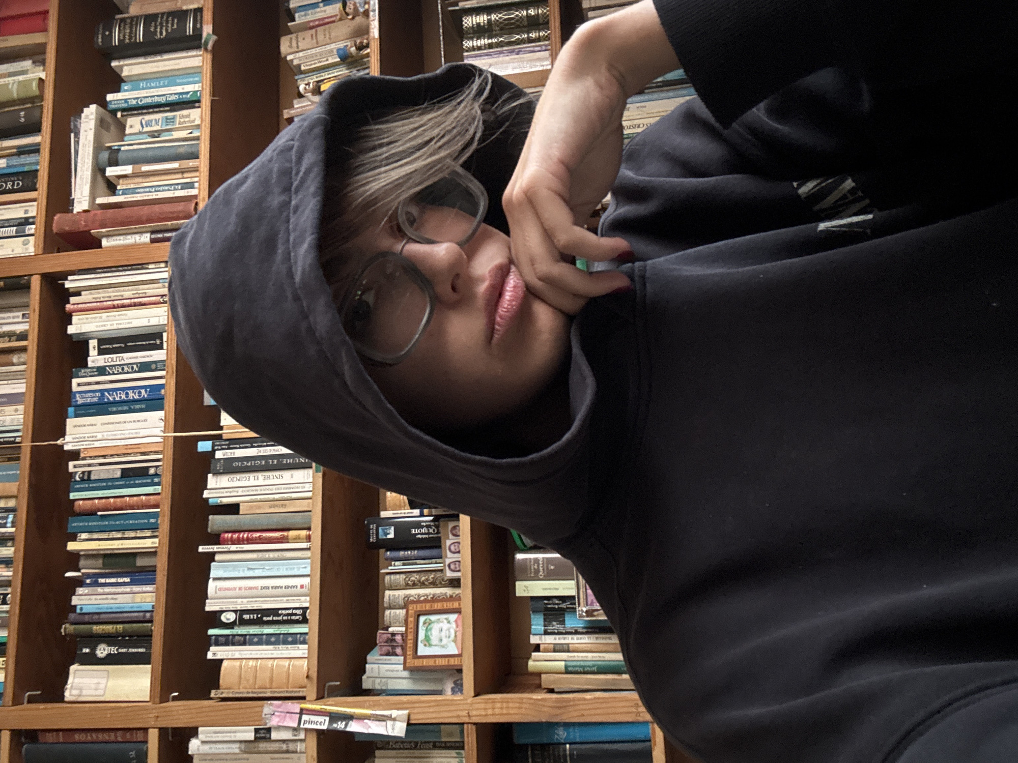 A person with light hair, wearing glasses and a black hoodie, is positioned in front of a bookshelf filled with books. The books appear to be a mix of literary works and academic texts in English and Spanish. Visible titles include "Hamlet," "The Canterbury Tales," "Lolita" (by Nabokov), "The Basic Kafka," "Aztec," and various titles in Spanish, such as "Sinúhe, el egipcio". There is also a small framed picture among the books. The person's face is turned towards the viewer, and they are resting their chin on their hand. The location is not explicitly identifiable as Mexico City; the scene appears to be indoors, likely a library or personal study.
