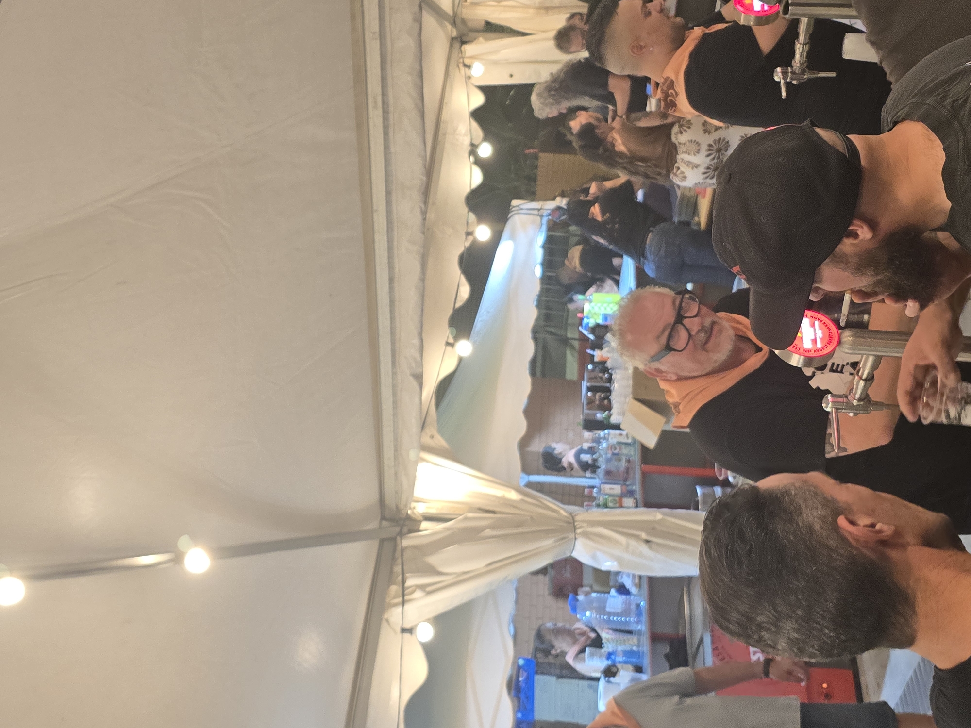 This image depicts an evening social gathering under white canopy tents in Cornellà De Llobregat, Spain. The scene is illuminated by multiple strings of bare bulb lights suspended across the tent's underside. In the foreground, three men are visible. On the far left, a man with short, dark, greying hair is seen from behind, wearing a dark t-shirt. To his right, an older man with white hair and a white beard, wearing glasses and an orange scarf, is smiling and looking right while standing behind a bar. His left hand is near a metallic beer tap system. To his right, a man with a dark beard, wearing a black baseball cap and a dark t-shirt, is seen from behind, facing the beer taps, possibly operating one or waiting for a drink. Further right, a man with dark hair, a faded haircut, and an orange scarf, wearing a dark t-shirt, also stands behind a bar with a visible beer tap.
In the midground and background, additional individuals are present. On the left, a woman with dark hair is partially visible behind a counter, where several large plastic water bottles and other items are arranged. Towards the center-right and background, multiple people are standing behind various bar setups containing an assortment of liquor bottles on shelves, indicating multiple serving stations. One woman in the mid-background wears a white skirt with a dark pattern, and another woman further back has long grey, curly hair. The background features a brick wall behind the bar areas. The ground appears to be a paved or concrete surface. The atmosphere is that of a bustling outdoor event or festival.