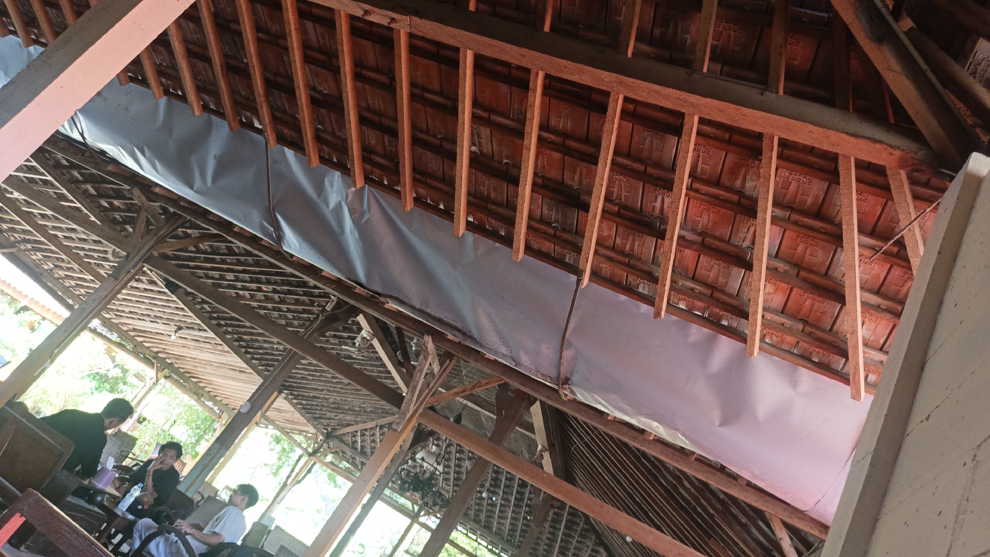 The image shows the interior of an open-air pavilion with a wooden roof structure covered with terracotta tiles. A light gray waterproof membrane is visible beneath the tiles, covering a section of the roof. Sunlight streams in from the sides, illuminating the wooden beams and rafters. In the foreground, three young men are seated at tables, engaged in conversation. One man, wearing a black shirt, is facing away from the camera. Another man, in a black shirt, is looking towards the camera. The third man, wearing a light-colored t-shirt, is also looking towards the camera. The scene appears to be a casual gathering in a tropical or sub-tropical environment, possibly a cafe or restaurant in Ngembalrejo, Indonesia.