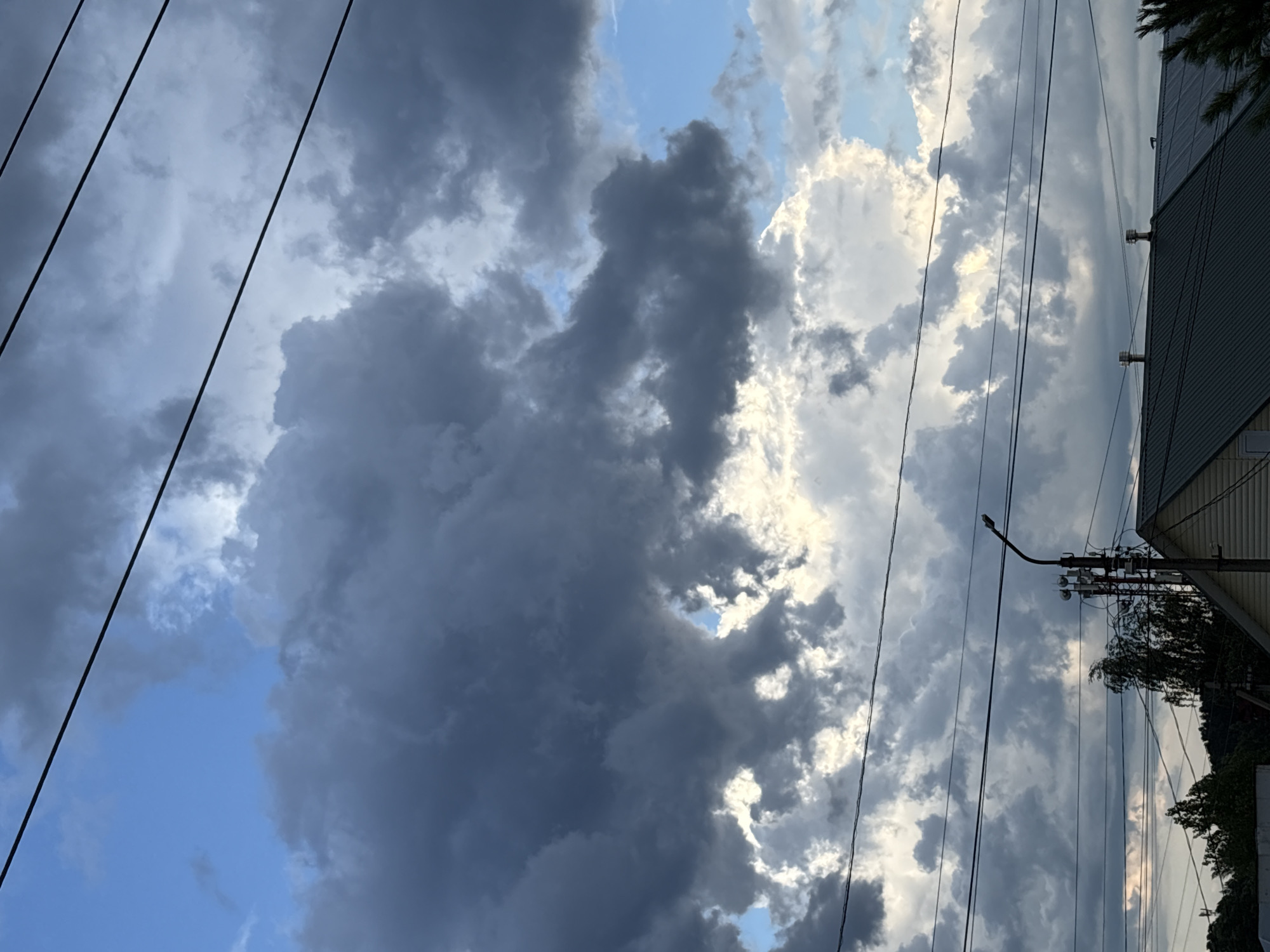 The image depicts a cloudy sky with dramatic, dark storm clouds interspersed with patches of bright sunlight. Several power lines crisscross the frame, creating a linear pattern against the sky. In the lower portion of the image, the rooftops of several buildings are visible, along with trees and a street lamp. The scene appears to be a residential area. The location is specified as Прикамский, Russia.