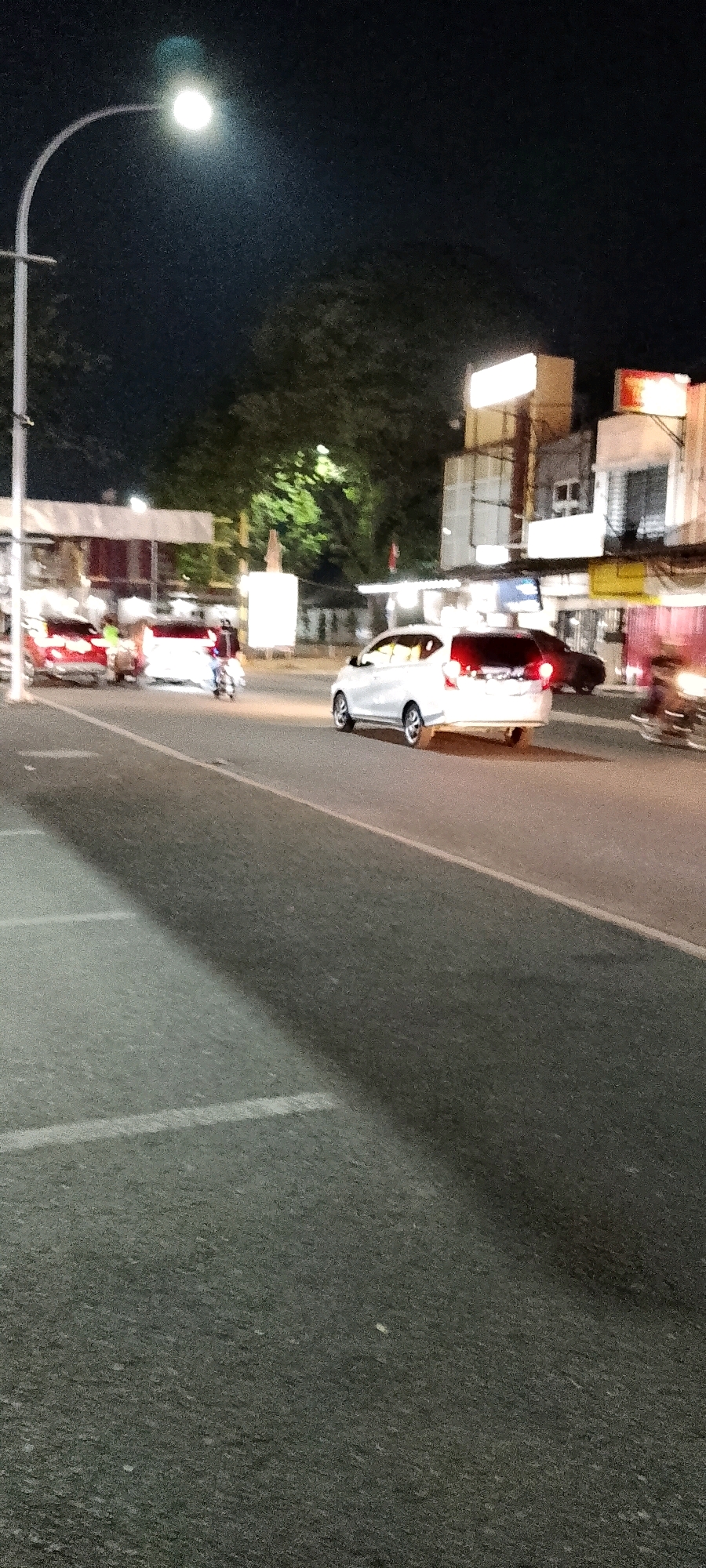 This is a nighttime street scene in Cileungsi, Indonesia. A white car is driving on a paved road. The car's brake lights are illuminated. On the left side of the road, there are parking lines visible on the asphalt. In the background, there are buildings with illuminated signs. Trees can be seen behind the buildings. A street light with a bright bulb is positioned on the left side of the frame. Other vehicles, including a red car and a motorcycle, are also present on the road, some with motion blur indicating movement. The overall lighting suggests it is after dark.