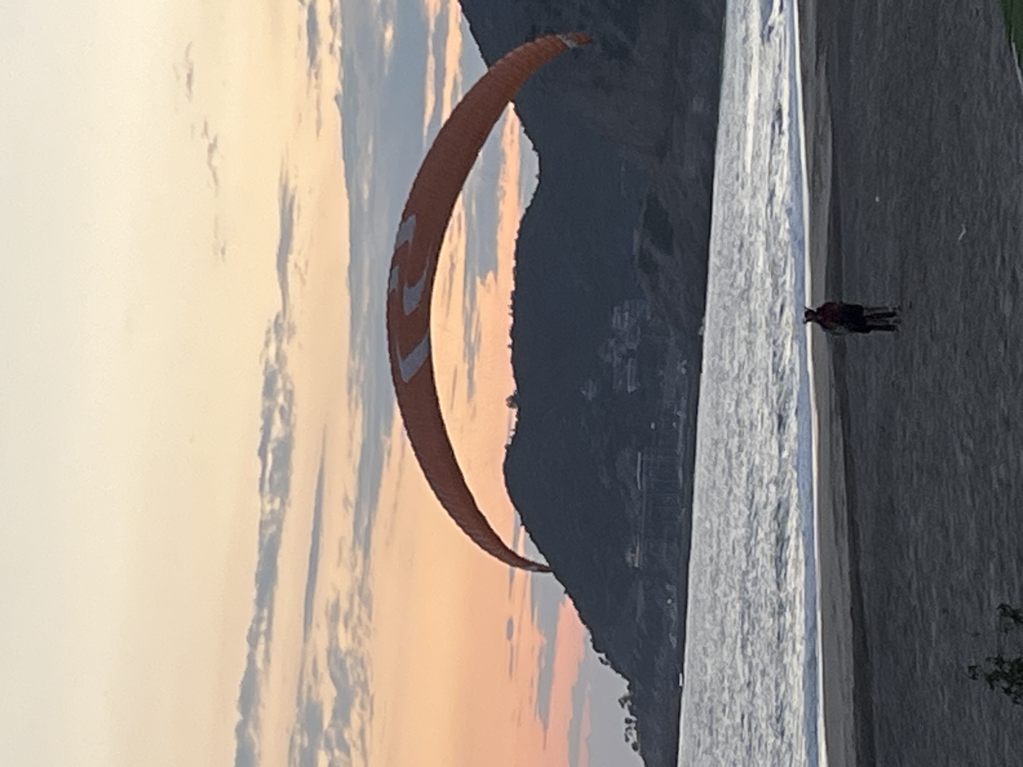 The image depicts a scene at Kleopatra Beach in Alanya, Turkey. A large, orange and white paragliding parachute is prominently featured against a backdrop of a mountain and a sky with hues of orange and blue indicating either sunrise or sunset. The Mediterranean Sea is visible, and two indistinct figures are standing on the beach.