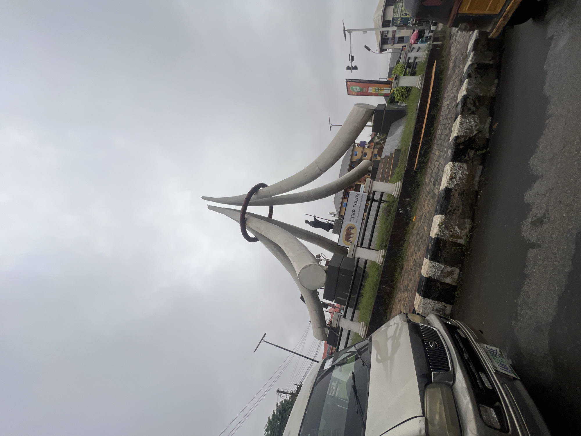 The image shows a large monument in the city of Onitsha, Nigeria, on an overcast day. The monument, situated in a traffic circle, consists of several massive, curved white structures that converge at the top, resembling elephant tusks. A statue of a person holding a staff stands at the base of the monument. A sign for "TIGER FOODS" is visible on a pedestal. In the foreground, the front of a silver minivan is captured, parked on a wet asphalt road. The minivan has a license plate that reads "AAH 739HE". Adjacent to the road is a curb made of alternating black and white painted concrete blocks, with some moss and dirt accumulation. Beyond the monument, buildings and traffic lights can be seen. To the right, part of a yellow and black auto-rickshaw is visible. The sky is uniformly gray, indicating cloudy weather, and the ground appears damp, suggesting recent rain.