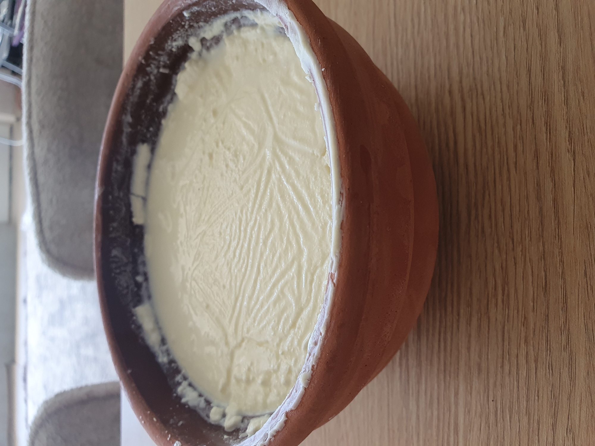 A close-up, eye-level shot shows a clay pot filled with a creamy, white substance, possibly yogurt or curd, resting on a light brown wooden surface. The texture of the contents within the pot is slightly uneven, with visible lines and swirls, suggesting it might have been stirred or is in the process of setting. The rim of the clay pot has a slightly rough, unglazed appearance. In the background, out of focus, are two grey upholstered chairs and a light-colored surface, possibly a table or floor. The overall scene is indoors, with soft lighting.