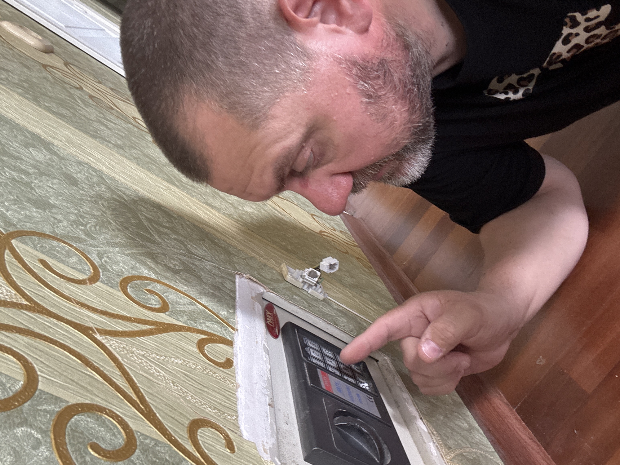 The image shows a close-up of a man's face and hand interacting with a small wall safe. The safe is partially embedded in a wall covered with patterned wallpaper. The man, with short brown hair and a beard, is looking at the safe's keypad, and his right index finger is touching one of the buttons. A white wire is attached to the safe's side. The wall safe appears to be a model from the brand ONYX. The surroundings suggest an indoor environment with a wooden floor and baseboard. The overall scene depicts an attempt to access or operate the safe. There is no information to suggest this scene is located in the city of Krasnodar, Russia.