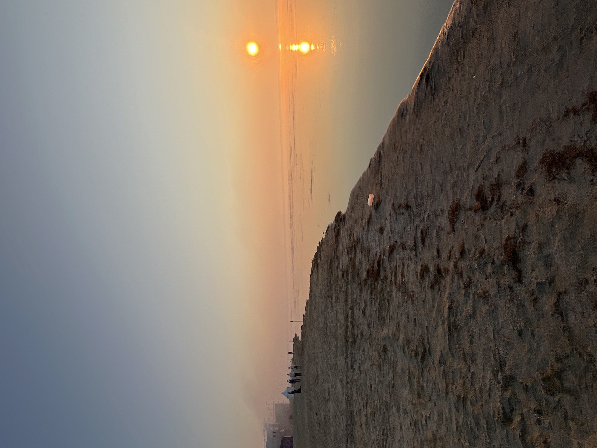 In Surakovo, Russia, at sunrise, the sun casts a warm orange glow over a beach and the calm sea. The sky transitions from a pale blue at the top to a soft orange near the horizon. Along the sandy shore, several people are gathered, some standing near tents and what appears to be a stage structure in the distance. The water reflects the bright sun, creating a shimmering path of light. A few pieces of litter are visible on the sand.