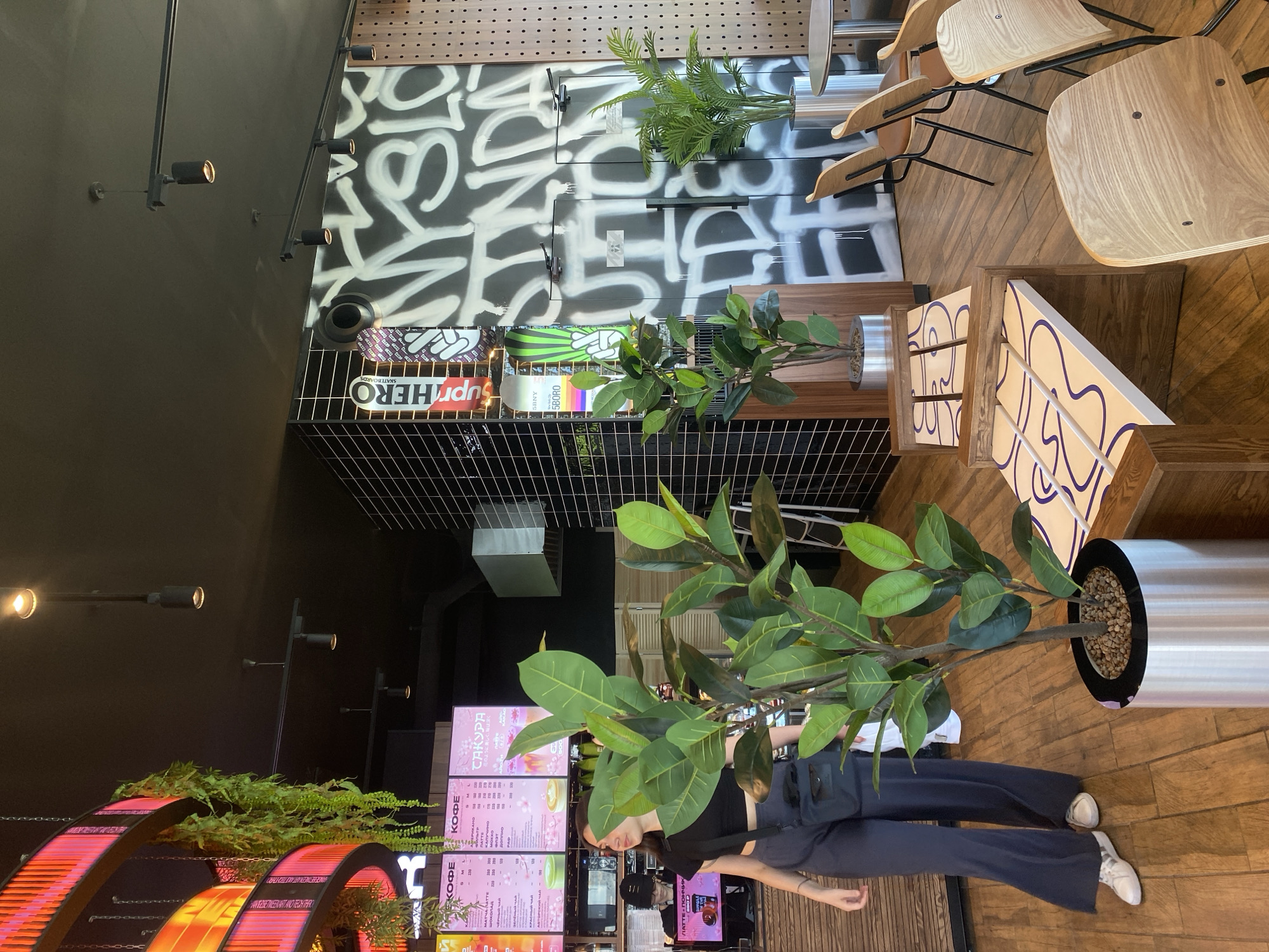 The image depicts the interior of a modern cafe. In the foreground, a young woman wearing a black crop top and dark wide-leg pants is standing and looking to the left, with a black crossbody bag. To her right, a large potted ficus plant stands in a silver cylindrical planter. The floor is made of wooden planks.
In the background, a wall is covered with graffiti. To the left of the graffiti, there is a menu board with Cyrillic text and images of drinks. Further left, a circular structure with pink and orange lighting hangs from the ceiling, with artificial green vines draped from it. Various light fixtures are visible on the dark ceiling.
To the right of the graffiti-covered wall, there are several modern wooden chairs with black metal frames arranged around tables. A smaller potted palm tree is also visible. The cafe appears to be clean and contemporary, with a mix of urban art and natural elements. The overall atmosphere is casual and trendy. Based on the Cyrillic text visible on the menu board, it is plausible this establishment is located in Perm, Russia.