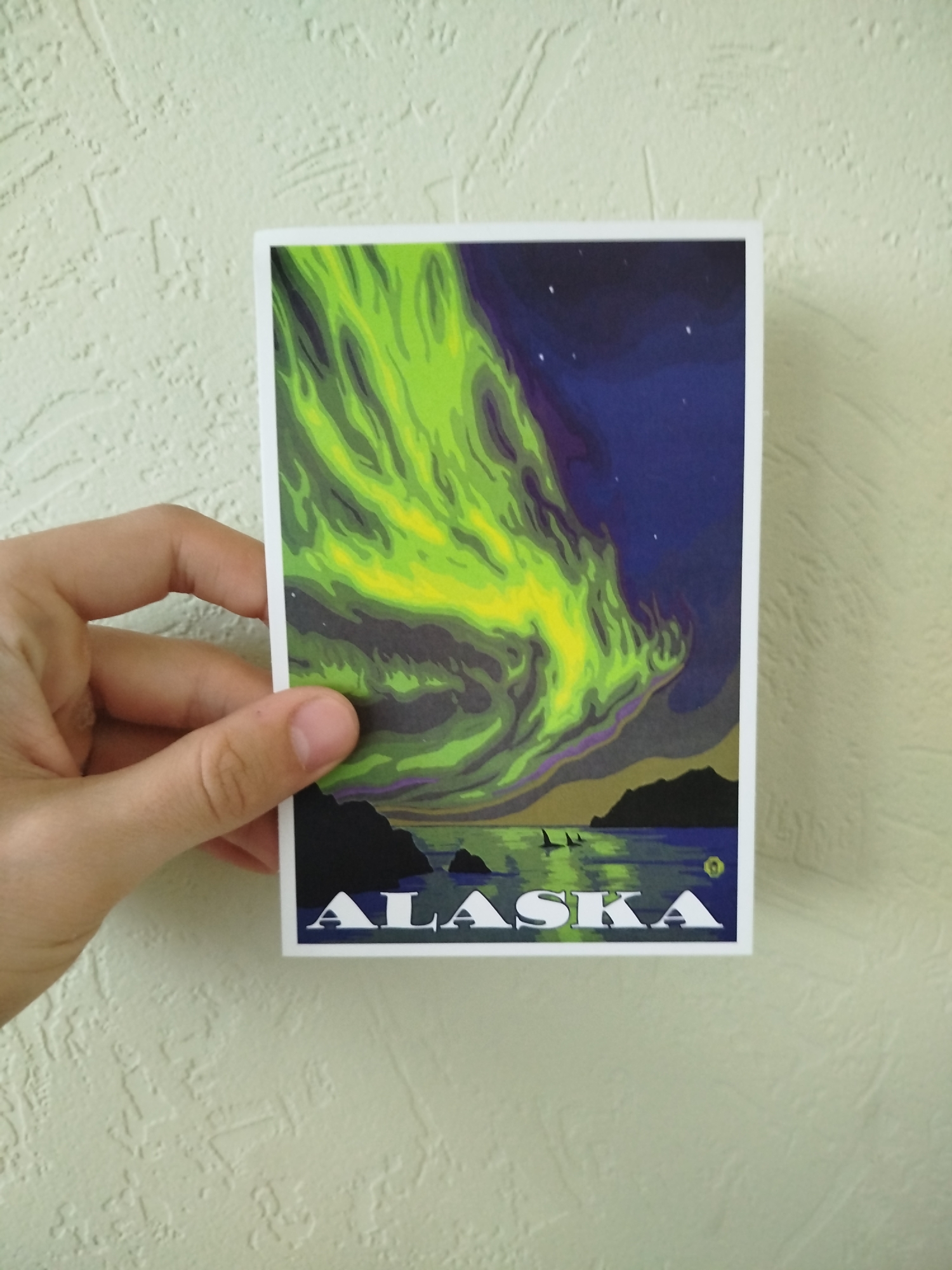 A postcard with a stylized illustration of the Alaskan Aurora Borealis is being held by a hand against a textured beige wall. The artwork depicts a vibrant green and yellow aurora swirling above a dark landscape with water and silhouetted landmasses. The word "ALASKA" is printed in bold white letters at the bottom of the postcard. There are no people visible besides the hand holding the postcard. The environment consists of the beige wall and the postcard. There is no indication of the location being Varna, Bulgaria.