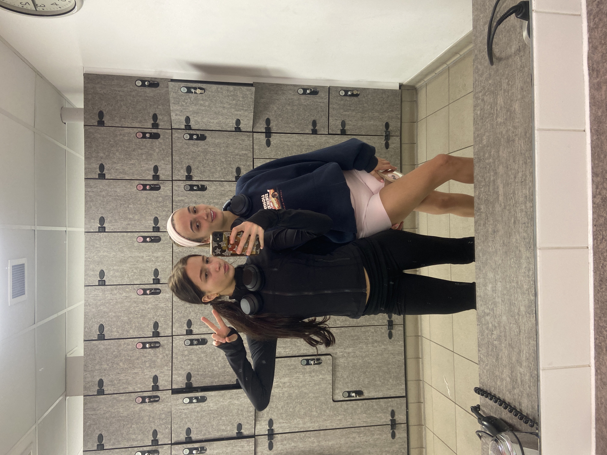Two young women are posing for a selfie in what appears to be a locker room or changing area. The person in the foreground is wearing black athletic apparel, including leggings and a zip-up jacket, and has a ponytail. She is holding up a peace sign with her left hand. Around her neck, she has black over-ear headphones. The other person is wearing a headband, a dark blue sweatshirt, and light pink shorts. She is holding the phone, taking the photo. Both have a casual, relaxed posture.
Behind the women are multiple rows of grey lockers. The floor is tiled in light-colored squares. Part of a mirror is visible on the right side of the image, reflecting the scene. There's a hair dryer with its cord visible. Above, a clock is on the wall.
Given the context and style, it’s plausible this locker room is located within a sports facility or gym in Mole Valley, United Kingdom.