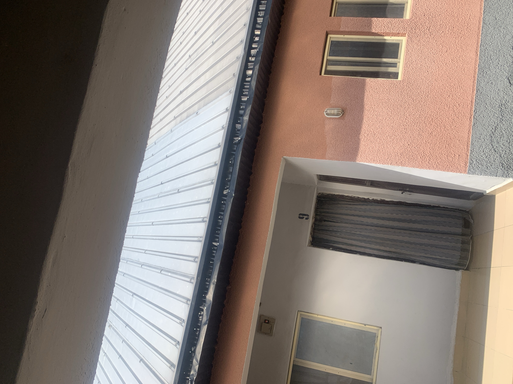 The image shows an exterior view of a building with a corrugated metal roof. The building has a light peach-colored textured wall with a gray lower section. There are several windows visible, some with light-colored frames and others with dark frames. One window on the left appears to have a screen. To the left of a window, there is a light switch. A door with a dark curtain is visible on the left side of the building, with the number "9" painted above it. A light fixture is mounted on the peach-colored wall. The foreground is partially obscured by an overhang, casting a shadow. The scene appears to be in Jauro Yinu, Nigeria. There are no people visible in the image.