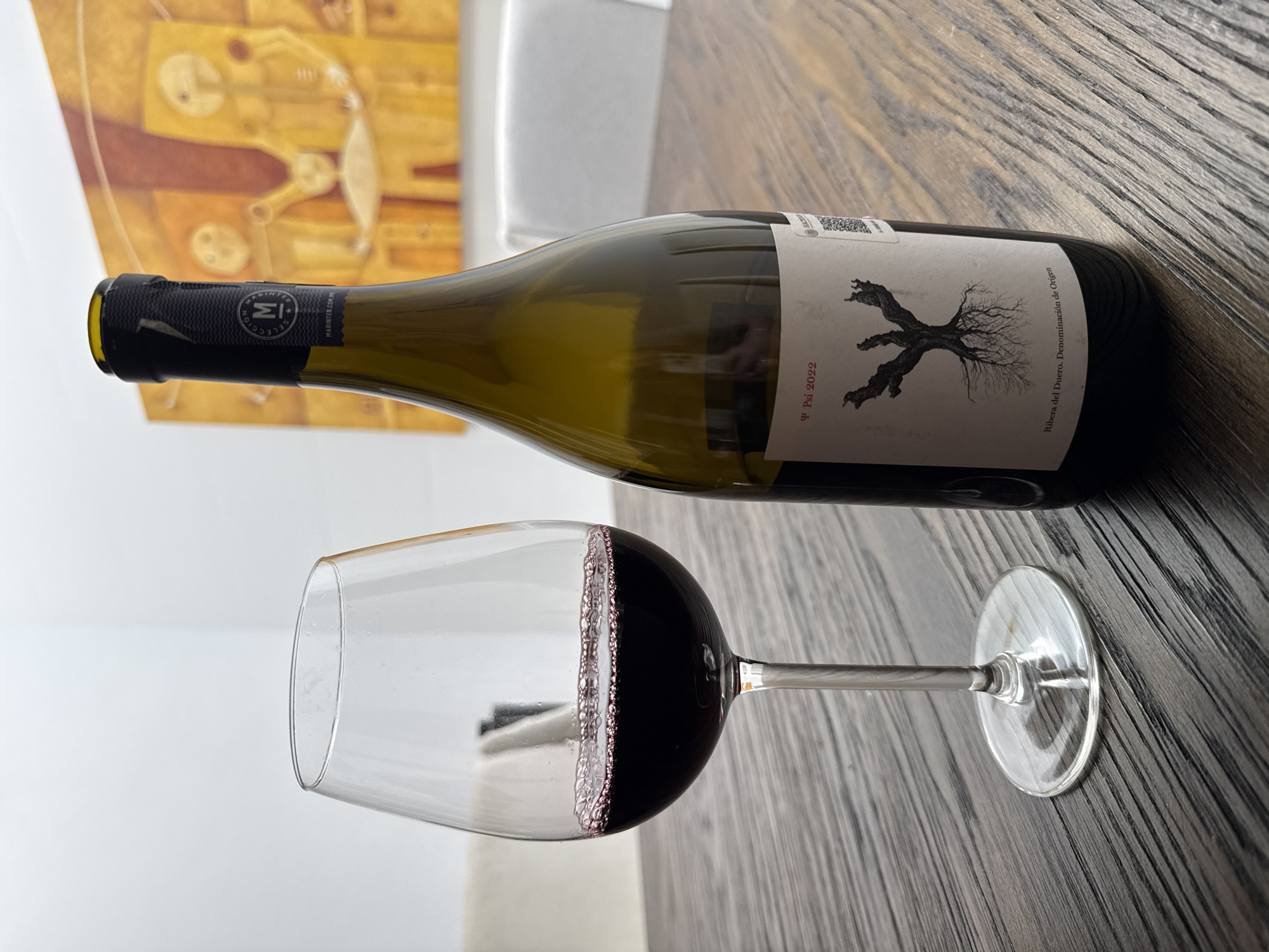 The image shows a wine bottle and a glass of red wine on a dark wooden surface. The bottle is green glass with a white label featuring a black tree-like design and the text "Ψ Psi 2022 Ribera del Duero. Denominación de Origen". The glass is partially filled with red wine, displaying bubbles along the wine-air interface. In the background, there is a painting with yellow and brown hues depicting abstract figures, and a white chair. The scene appears to be indoors. It is impossible to ascertain the location is in Huixquilucan de Degollado, Mexico, based on the visual information provided. There are no people in the frame.