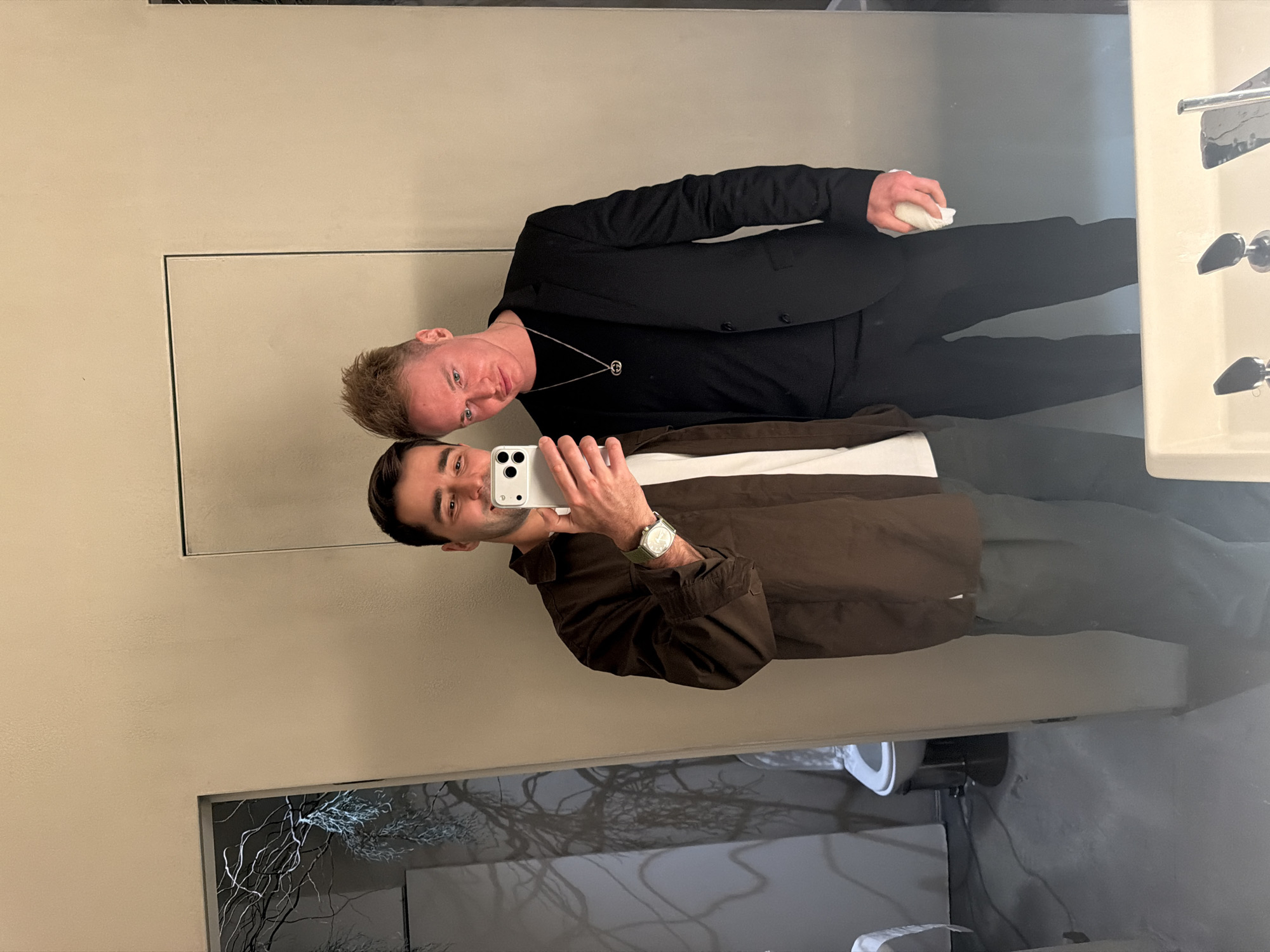 Two men are posing for a mirror selfie in a restroom. The man closest to the camera is holding a smartphone. He is wearing a brown jacket, a white shirt underneath, grey pants, and a watch. The other man is wearing a black suit, a black shirt, and a necklace. He is holding a white cloth. The restroom has a sink with a faucet and a mirror. There is a framed picture or display on the wall with a grey abstract tree design. It is not possible to confirm the location as Moscow, Russia from the image alone.