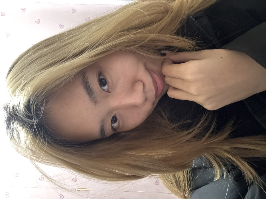 A medium-close-up portrait depicts an East Asian female subject indoors, within Shapingba District, China. The subject is positioned facing the camera with a slight head tilt to her left and a faint, closed-mouth smile. She has dark eyes and a light complexion. Her hair is long, appearing light brown or blonde with noticeable dark roots. Her left hand is placed under her chin, with fingers gently curved. She is wearing a dark-colored upper garment, possibly a jacket. The background is a light-colored wall, possibly off-white or light pink, adorned with scattered small pink heart-shaped decals. The specific address or type of building within Shapingba District cannot be determined, indicating a private or residential interior setting.