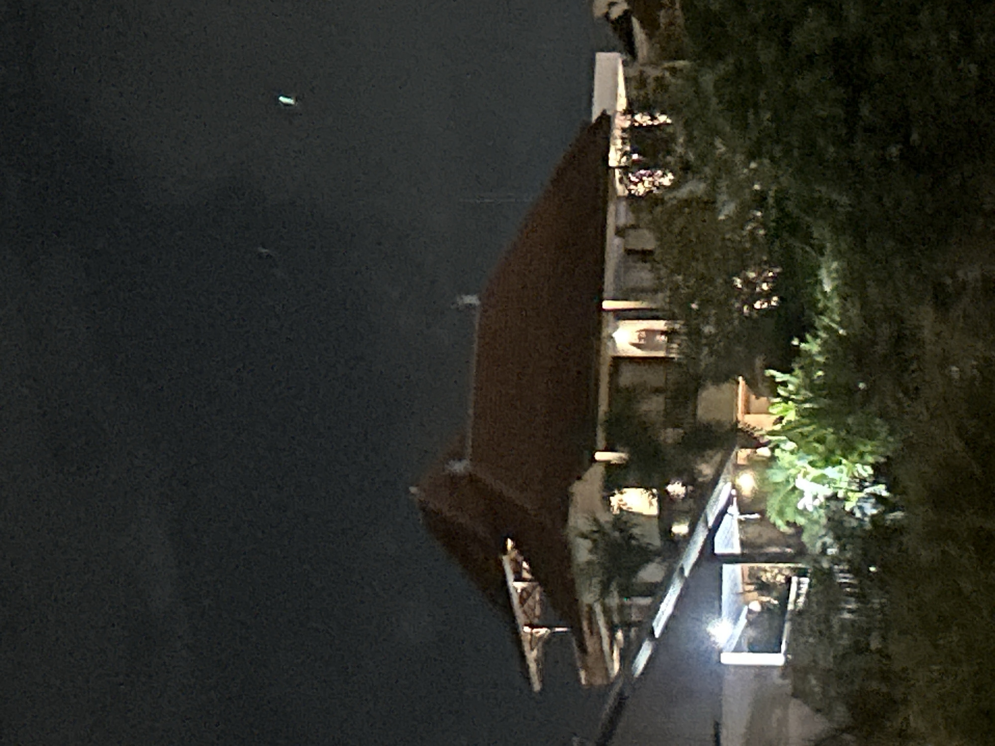 A medium shot image captured at night shows a building with a multi-level roof and red tiles. The building is illuminated from within by warm yellow lights, with some parts of the facade also lit. Some plants are visible in the foreground, partially obscuring the lower levels of the building. The sky above is dark, with a few faint lights visible. The scene appears to be in a residential or commercial area, possibly a hotel or resort, given the architectural style and landscaping. The location is identified as North Kuta, Indonesia.