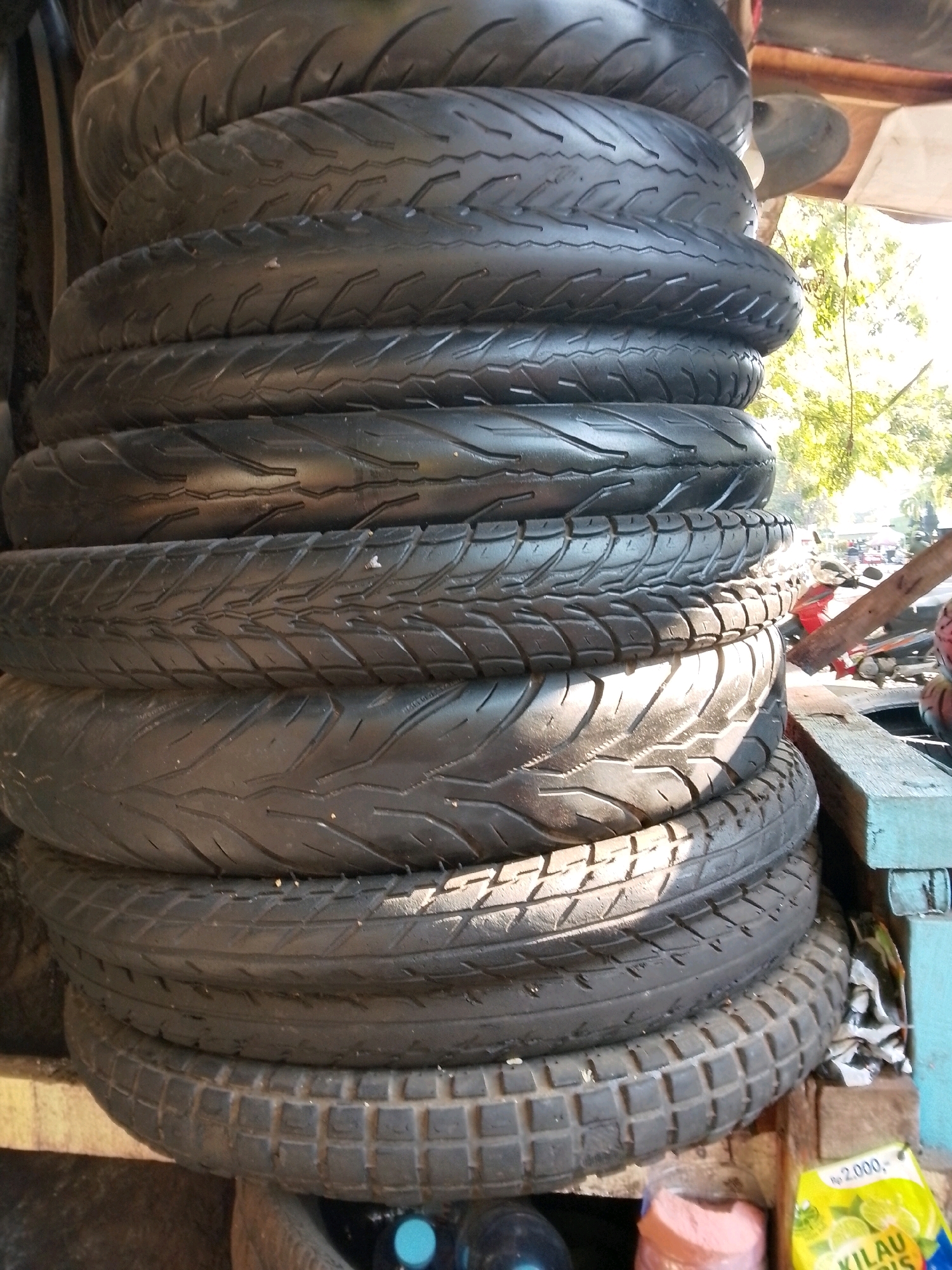 A stack of black rubber tires is displayed in an outdoor marketplace in Surabaya, Indonesia. The tires are of various sizes and tread patterns, with some appearing newer and others more worn. They are arranged in a leaning stack, with the largest at the bottom and tapering towards the top. The background reveals a bustling street scene with other market stalls, vehicles, and trees. The lighting suggests it is daytime.