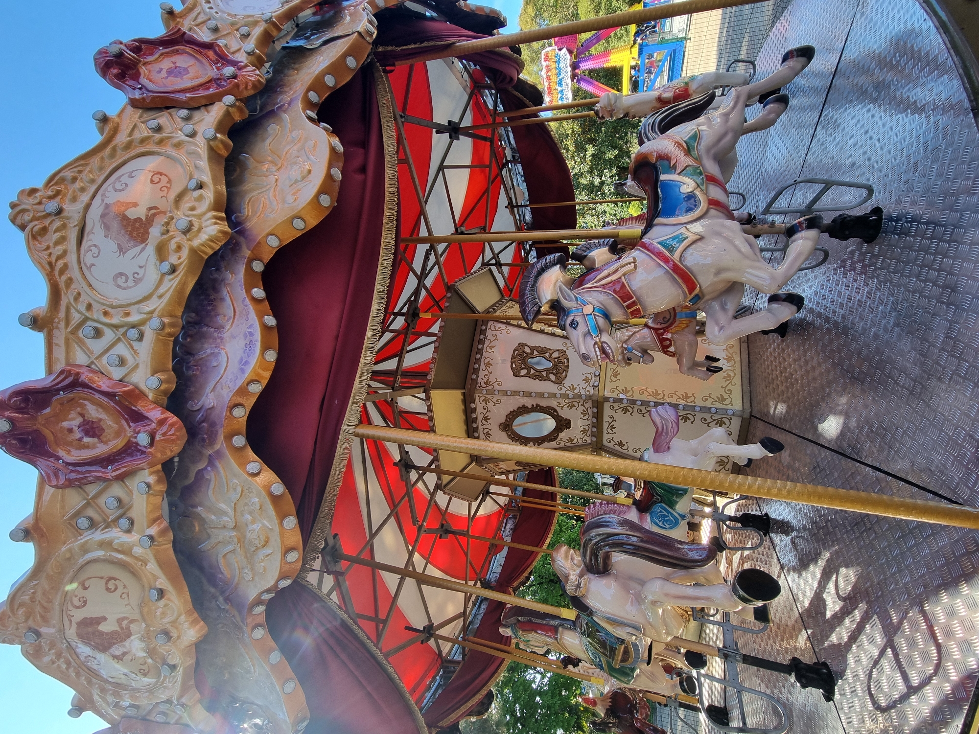 The image shows a carousel, a type of amusement ride, with several ornately decorated horses. The carousel is partially covered by a red and white striped canopy. The structure is detailed with painted panels and decorative trim. The background includes glimpses of trees and what appears to be another amusement ride, possibly a Ferris wheel, suggesting the setting is an amusement park or fairground. It is specified to be located in Sochi, Russia.
