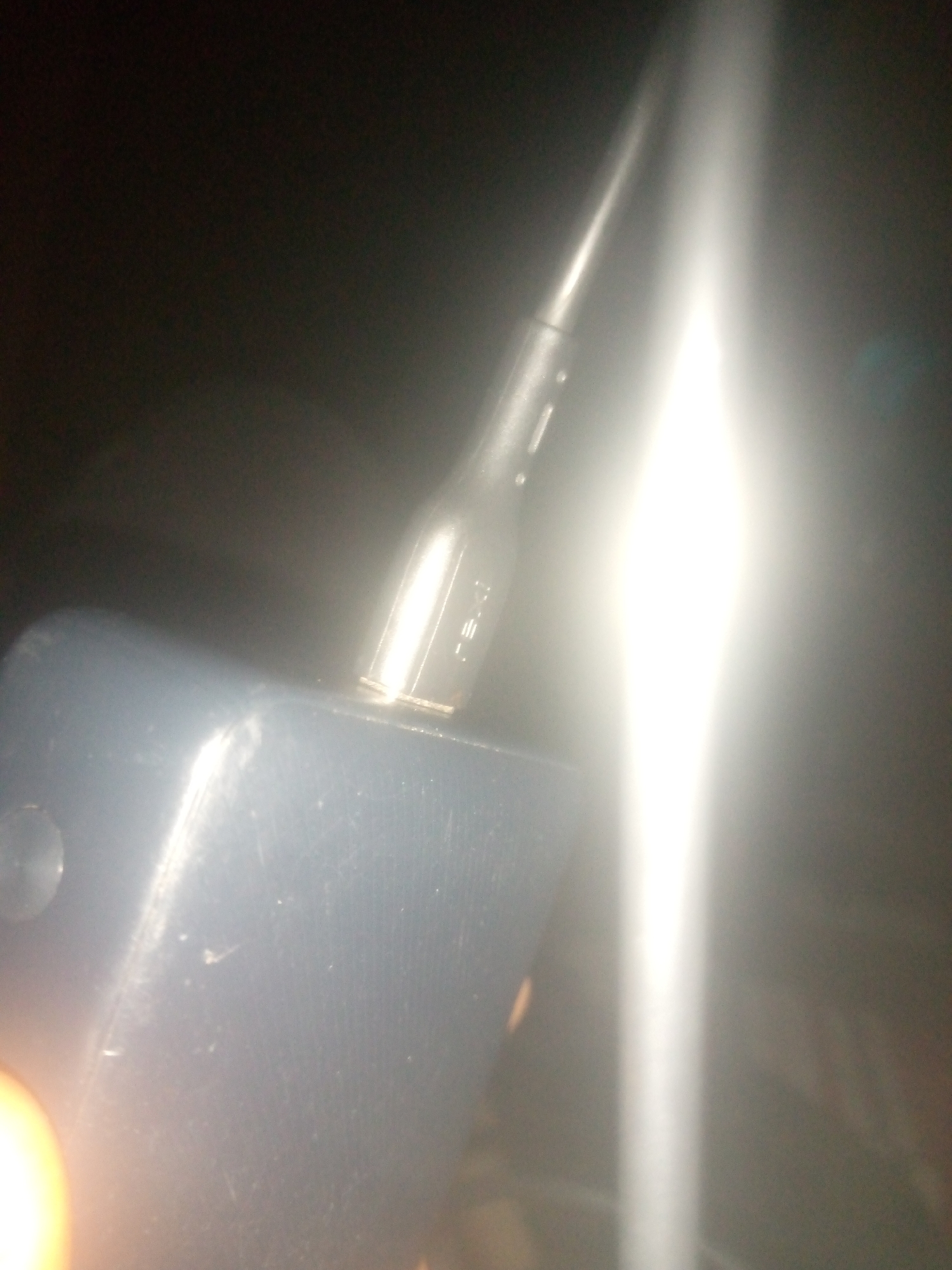 A close-up shot shows a black USB cable plugged into a dark blue device. The cable is inserted into a port on the device, and a bright light emanates from the area where the cable is connected, creating a strong glare that obscures details. The device appears to be a rectangular object with a matte finish. The background is dark and out of focus, with some light reflections visible on the device's surface.