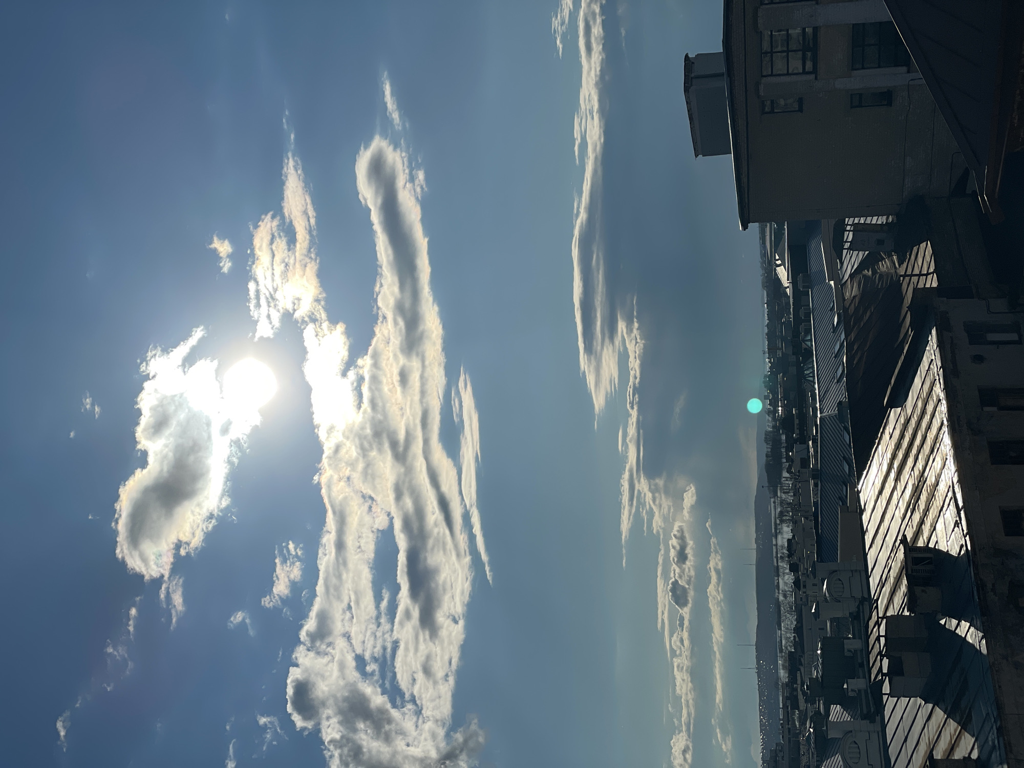 The image captures a vibrant daytime scene dominated by a vast blue sky with scattered clouds, viewed from an elevated perspective overlooking an urban environment.
The sun, positioned prominently on the left side, shines brightly through and around several clouds, creating a strong lens flare and illuminating the edges of the cloud formations. One particular cloud near the sun displays subtle iridescence, showing faint rainbow colors. Streaky, elongated clouds stretch across the upper and middle parts of the sky, contrasting with the deeper blue. A distinct bright green circular lens flare is visible towards the right side of the image.
In the lower half, a cityscape unfolds with numerous buildings of varying heights and architectural styles, suggesting an older, established urban area. The structures closest to the viewer appear to be multi-story buildings, possibly commercial or residential, with visible rooftops and air conditioning units. The sun casts strong shadows and highlights on the facades of these buildings, indicating the time of day. In the far distance, a hazy horizon reveals distant hills or mountains, adding a natural element to the urban landscape.
The overall impression is a clear, sunny day, likely midday or early afternoon, with good visibility, offering a wide panoramic view over the city towards the distant terrain. No specific activity or event is visible, and there is no discernible text within the image.