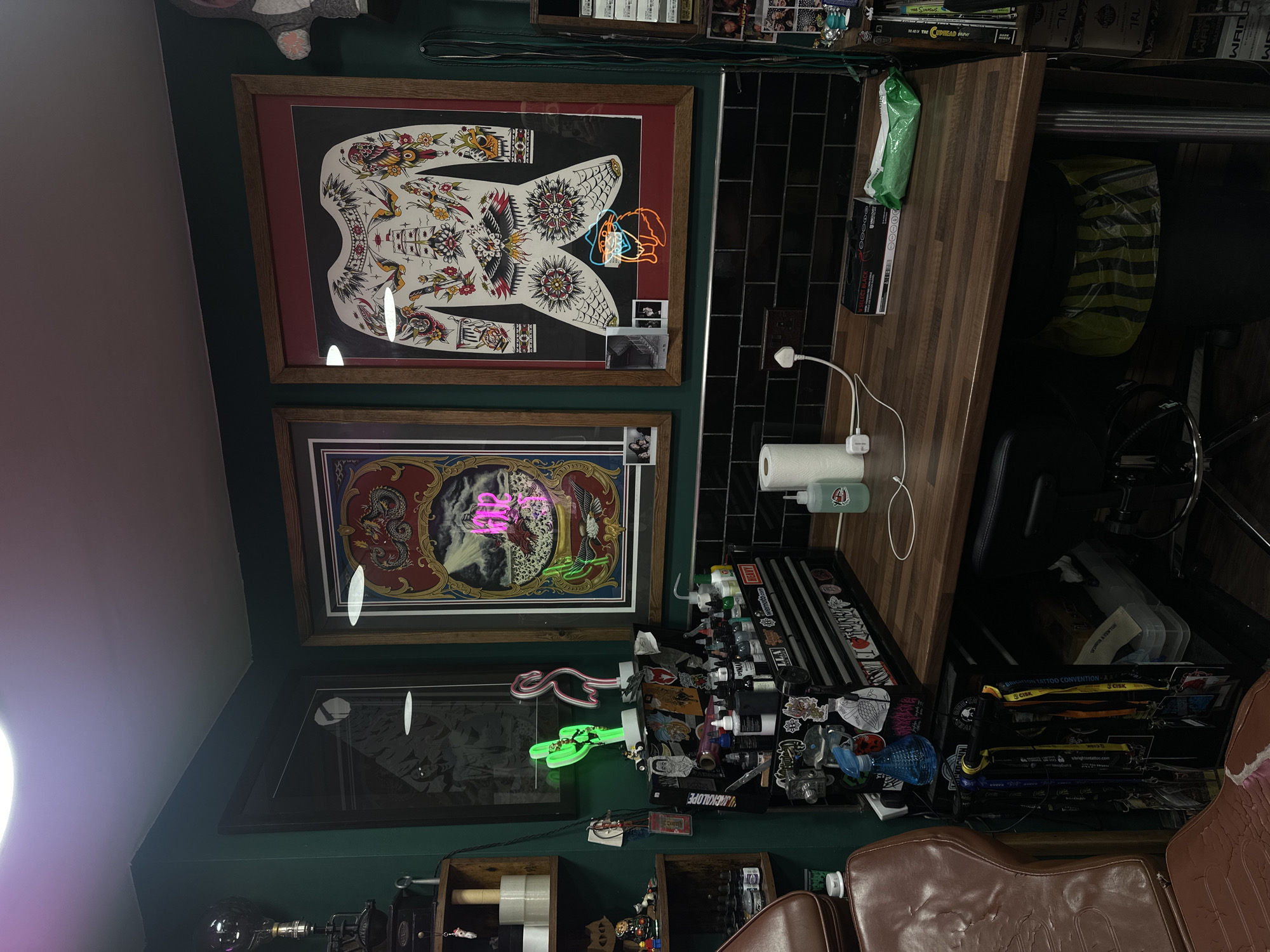 The image depicts a tattoo artist's workspace, likely in Brighton, UK, given the "BRIGHTON TATTOO CONVENTION" stickers and Brighton-related URL on lanyards. Two framed pieces of tattoo art adorn the wall above the workstation. A dark wood countertop sits above a black tile backsplash. Below this, a cluttered workspace features bottles of ink, medical supplies, and tattoo paraphernalia. A black adjustable chair and a worn, brown leather tattoo chair are visible. The room is dimly lit with green walls and accented with neon lights in the shapes of a flamingo and a cactus. Stickers and small objects are strewn around the desk.