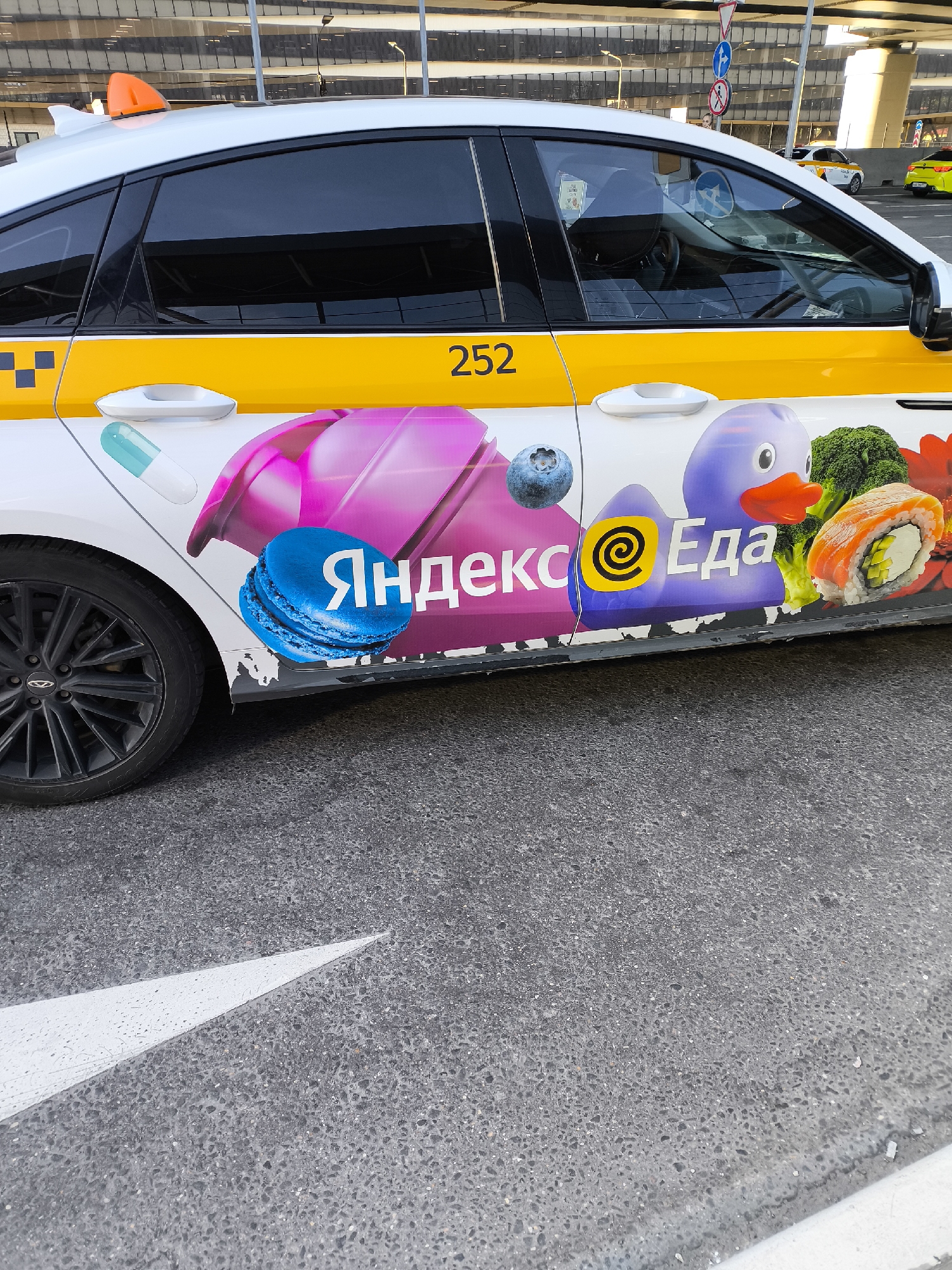 A white taxi sedan, identified by a yellow side stripe with the number "252" and an orange roof light, is parked on asphalt at Sheremetyevo International Airport (SVO) in Khimki, Russia. The vehicle's lower side features a colorful advertisement for "Yandex Eda" (Yandex Food), displaying a blue macaron with text, a blueberry, a purple cartoon duck, broccoli florets, a sushi roll, and a teal capsule against a pink abstract background. The car has black-tinted rear windows and a black multi-spoke alloy wheel on the front left. The asphalt ground in the foreground displays a white painted directional arrow. In the background, a large, modern airport terminal building with reflective glass facades is visible, along with other vehicles, including another white taxi and a yellow car, and various traffic signs.