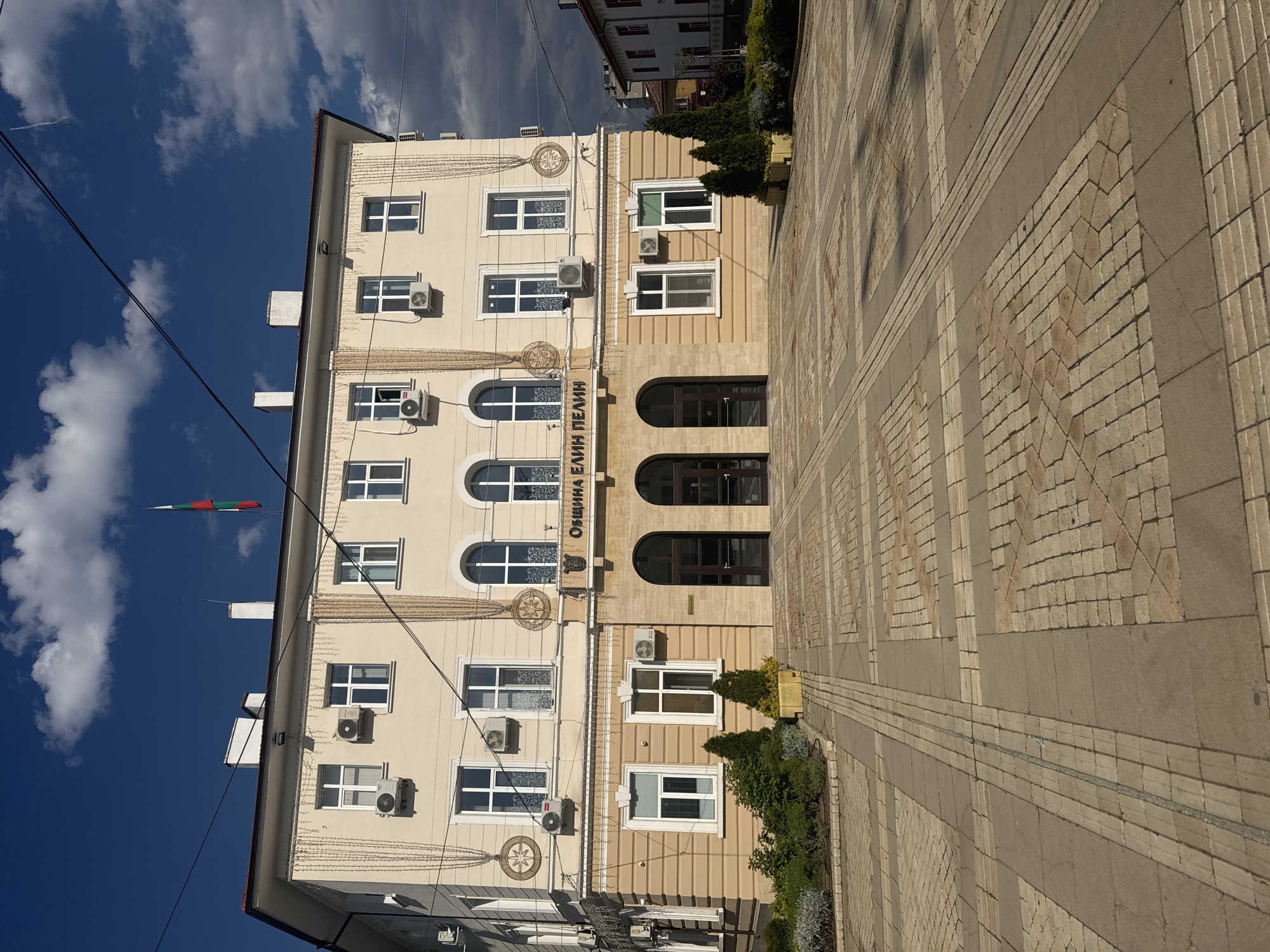 The image shows the municipal building of Elin Pelin, Bulgaria. The building is three stories tall, with a facade that combines light beige and tan colors. There are numerous windows, some rectangular and some arched, as well as air conditioning units attached to the building's exterior. A Bulgarian flag is visible. The ground in front of the building is paved with light brown patterned tiles. The sky is partly cloudy.