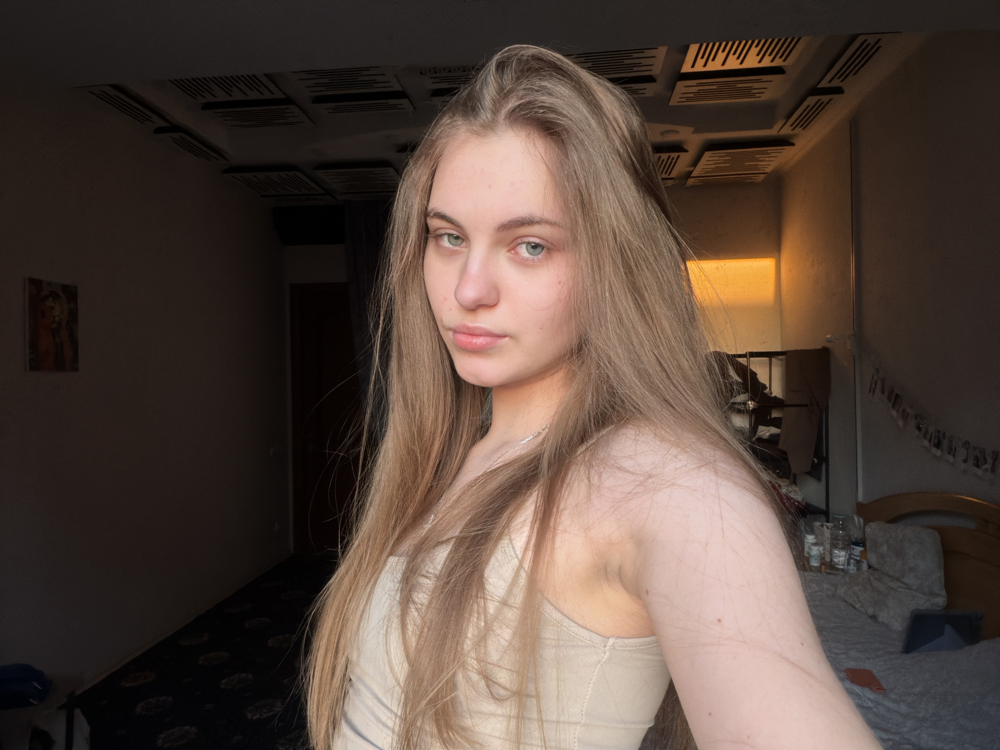 A young woman with long, blonde hair and light-colored eyes is pictured in a selfie taken in what appears to be a bedroom. She is wearing a beige tank top and a silver necklace. The room is dimly lit, with natural light streaming in from the right side of the frame, casting a warm glow. In the background, a bed is visible with pillows and a headboard. The ceiling has acoustic panels installed. To the left, a dark carpet with a pattern is visible on the floor, and a framed artwork hangs on the wall. The overall impression is a casual, indoor setting.