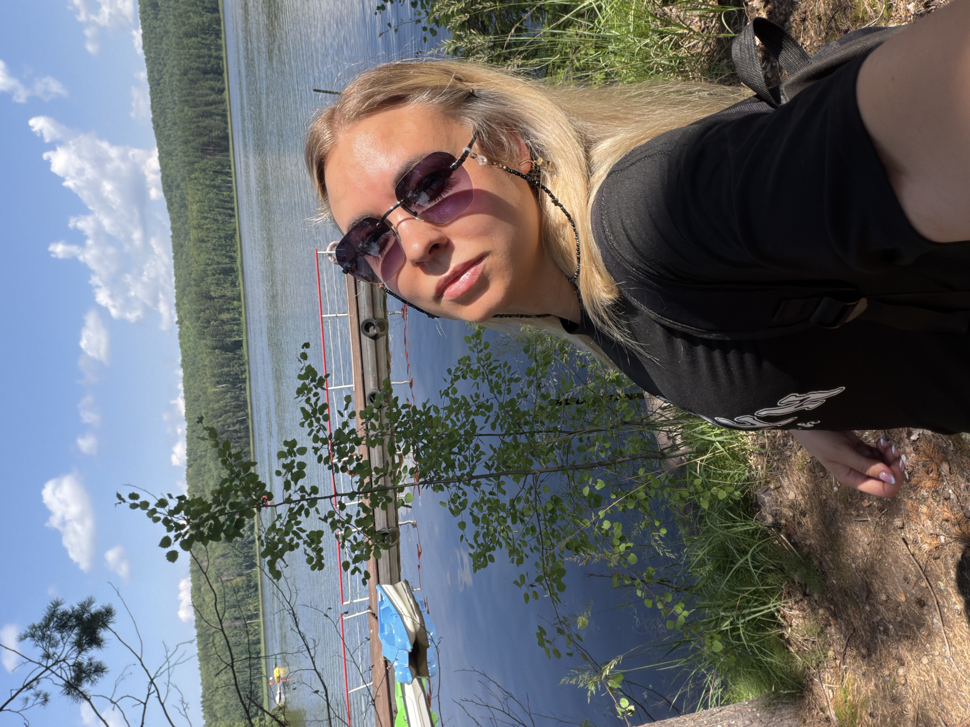A woman with blonde hair and sunglasses is taking a selfie by a lake. She is wearing a black t-shirt and a backpack. In the background, there is a wooden pier with two small boats, a forest, and a blue sky with white clouds. The location is the Kama River embankment in Chaykovsky, Russia.