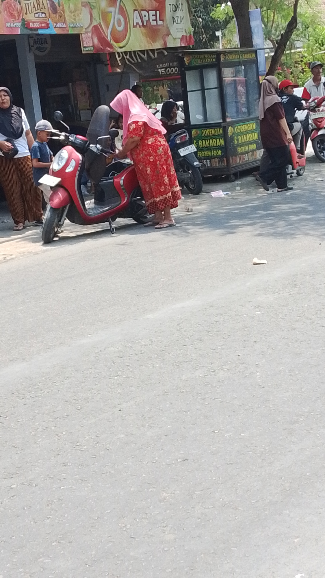 The image depicts a street scene in Mancar, Indonesia. In the foreground, a red scooter is parked. A woman in a red patterned dress and pink headscarf is attending to the scooter, her feet clad in flip-flops. To her left, a young boy in a blue shirt and a grey baseball cap stands near another parked motorcycle. Further back, a food cart is visible, with signage indicating "GORENGAN & BAKARAN" (Fried & Grilled) and "FROZEN FOOD". People are interacting around the food cart and the scooters, with some individuals on motorcycles and others walking. The environment appears to be a sunny day on a paved road, with trees and buildings in the background.