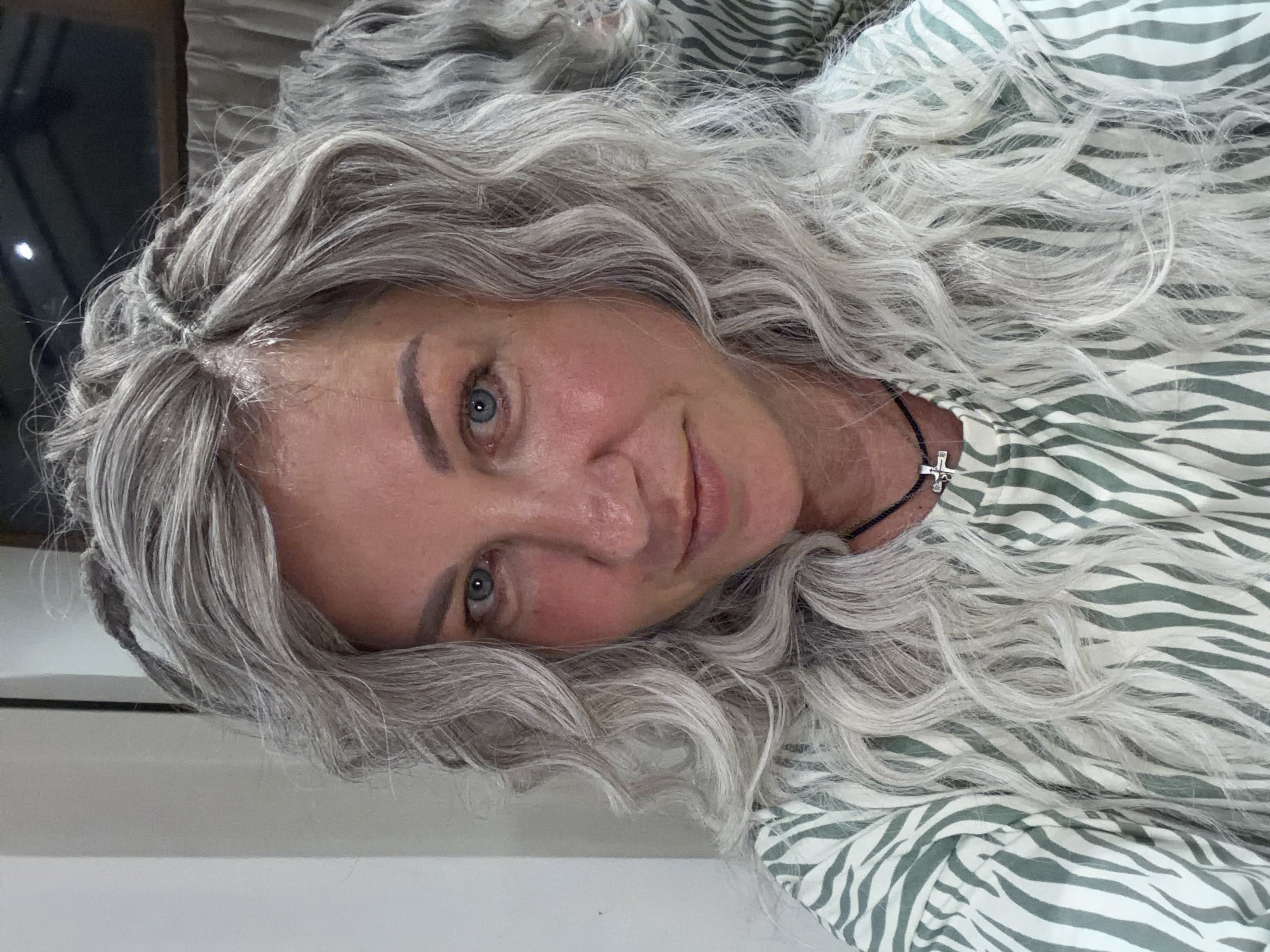 A photograph depicts a woman with light gray hair and fair skin, oriented at an angle such that her face is the focal point. Her hair appears to be wavy and voluminous, framing her face. Her eyes are light colored, and she has a slight smile. She is wearing a shirt with a green and white striped pattern. A thin black necklace with a small cross pendant is visible around her neck. The background is indistinct, with a hint of a curtain or window at the top left corner and a wall at the bottom. The setting is Unknown, Unknown.