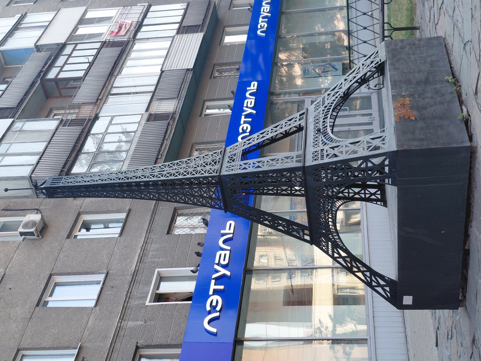 A miniature Eiffel Tower sculpture is positioned in front of a building with a blue sign that reads "Л'ЭТУАЛЬ". The sculpture is made of dark grey metal and stands on a dark grey rectangular base. Pigeons are perched on the blue sign. The building has multiple windows and balconies, and the facade is made of grey concrete. A paved walkway is in the foreground, and grass and a bench are visible in the background.
