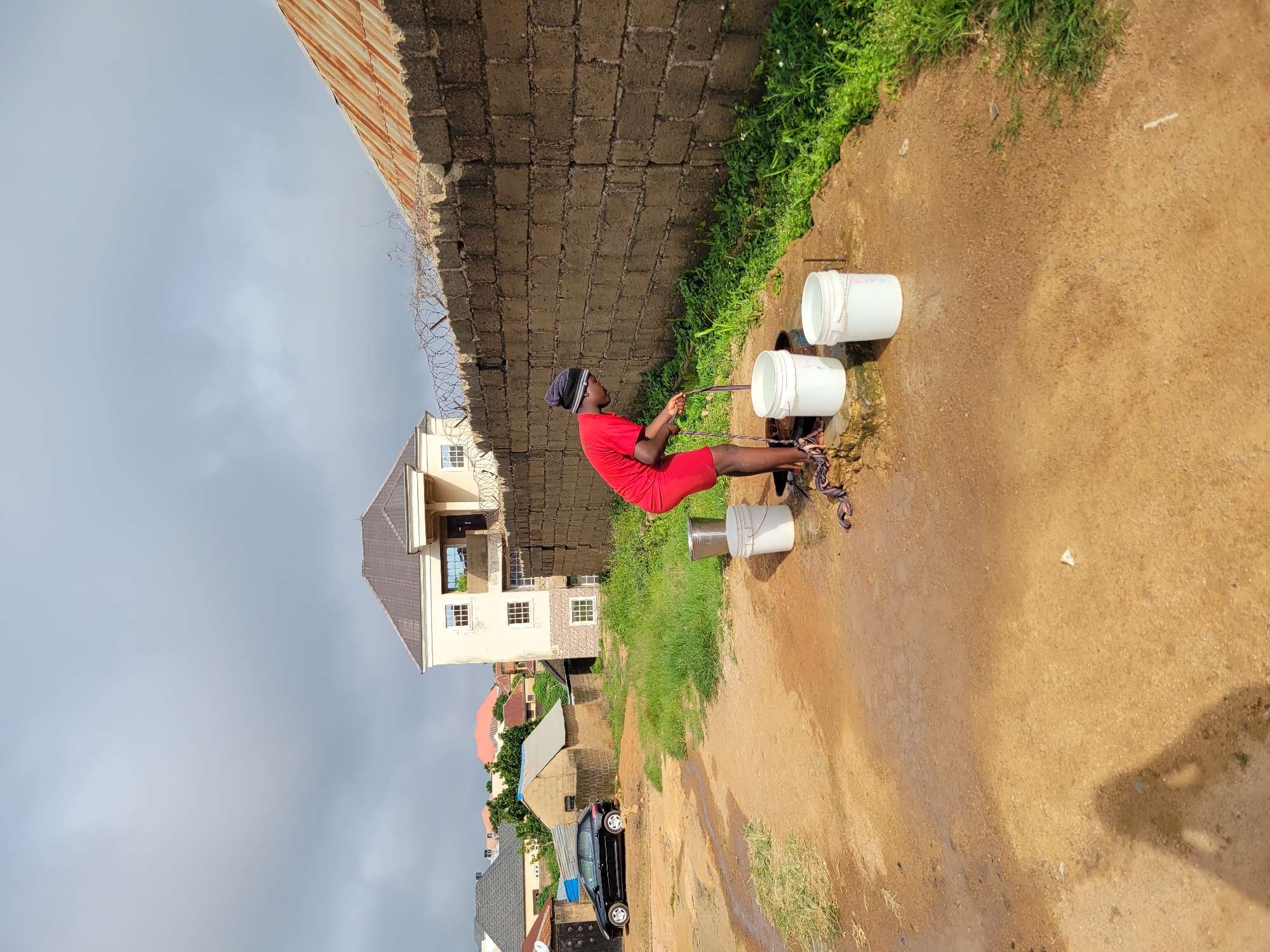 In Jos, Nigeria, a young person wearing a red t-shirt and shorts is drawing water from a well. They are using a purple rope to lower a bucket into the well, and another person's shadow is visible on the ground, suggesting they are taking the photograph. Three white buckets are placed near the well, one of which is partially filled with water. The scene is set in a dusty, outdoor area with overgrown green grass. In the background, there are several buildings, including a two-story structure with a light-colored facade and a rusted corrugated metal roof, and a black car parked on a dirt road. A brick wall with barbed wire runs along the right side of the image. The sky is overcast with clouds.