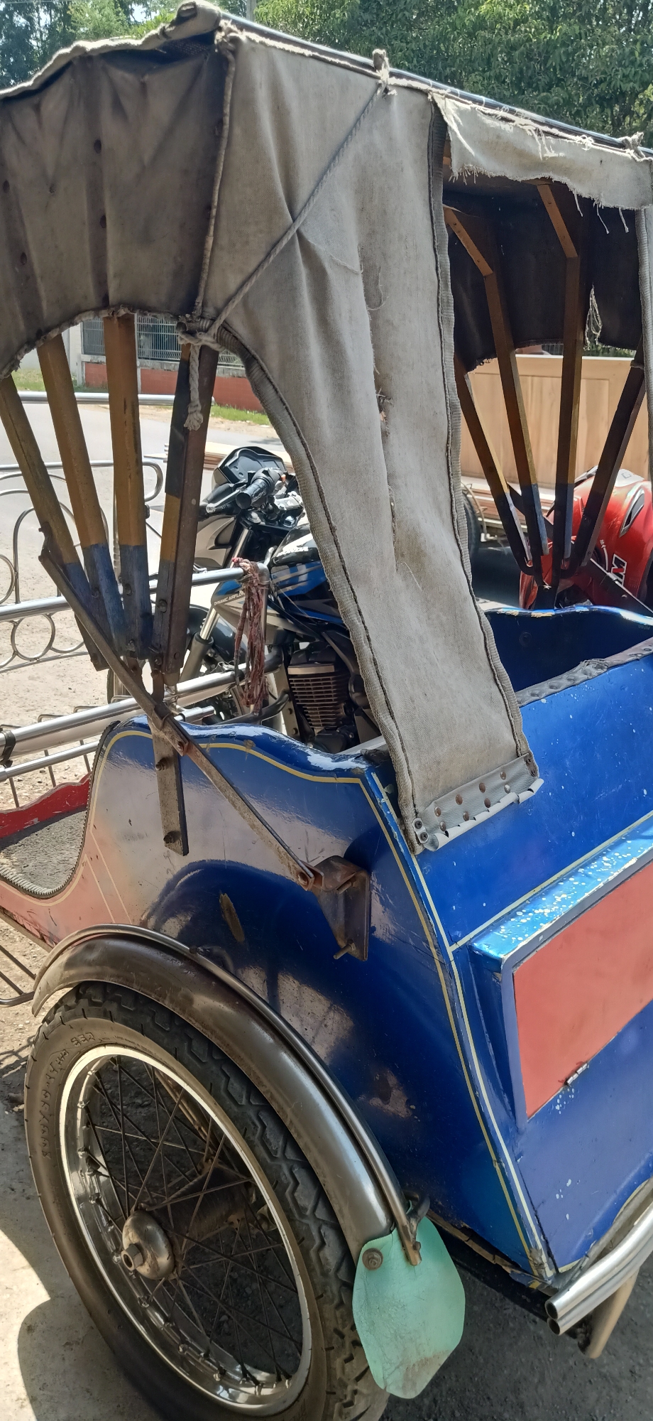 A three-wheeled motorized rickshaw, also known as a becak motor, is parked outdoors in Palohlada, Indonesia. The vehicle features a blue body with red and yellow accents. A weathered, light-colored canvas canopy covers the passenger compartment. The rickshaw is equipped with a spoked wheel at the rear. Behind the canopy, a motorcycle engine and handlebars are visible, indicating the vehicle's powertrain. A red helmet is also partially visible in the rear passenger area. The scene appears to be a street or roadside location, with trees and buildings in the background. The lighting suggests it is daytime.