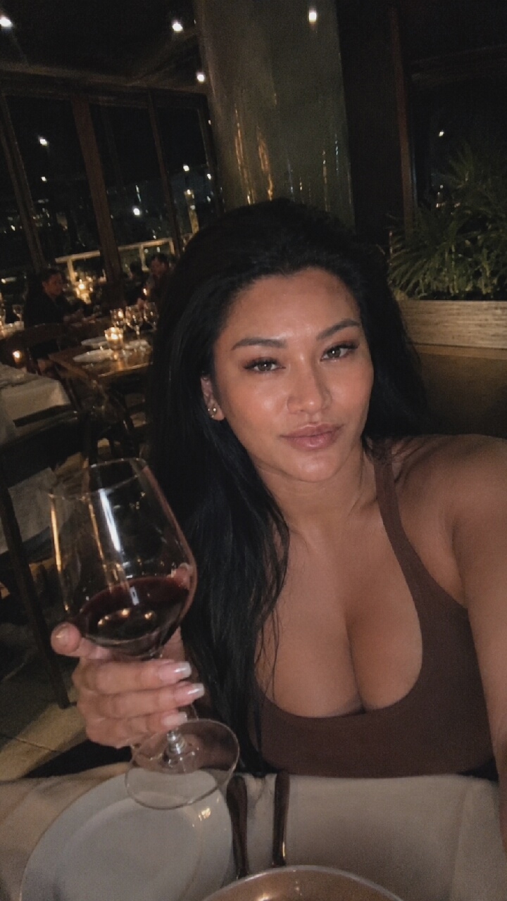 A woman with long, dark hair and light makeup is taking a selfie in a dimly lit restaurant. She is holding a glass of red wine in her left hand and looking at the camera with a slight smile. She is wearing a brown tank top. The background shows tables set for dinner, with people seated at them and city lights visible through large windows. The scene suggests a sophisticated dining experience in a city at night. There are no specific details that would indicate the location is Moscow, Russia.