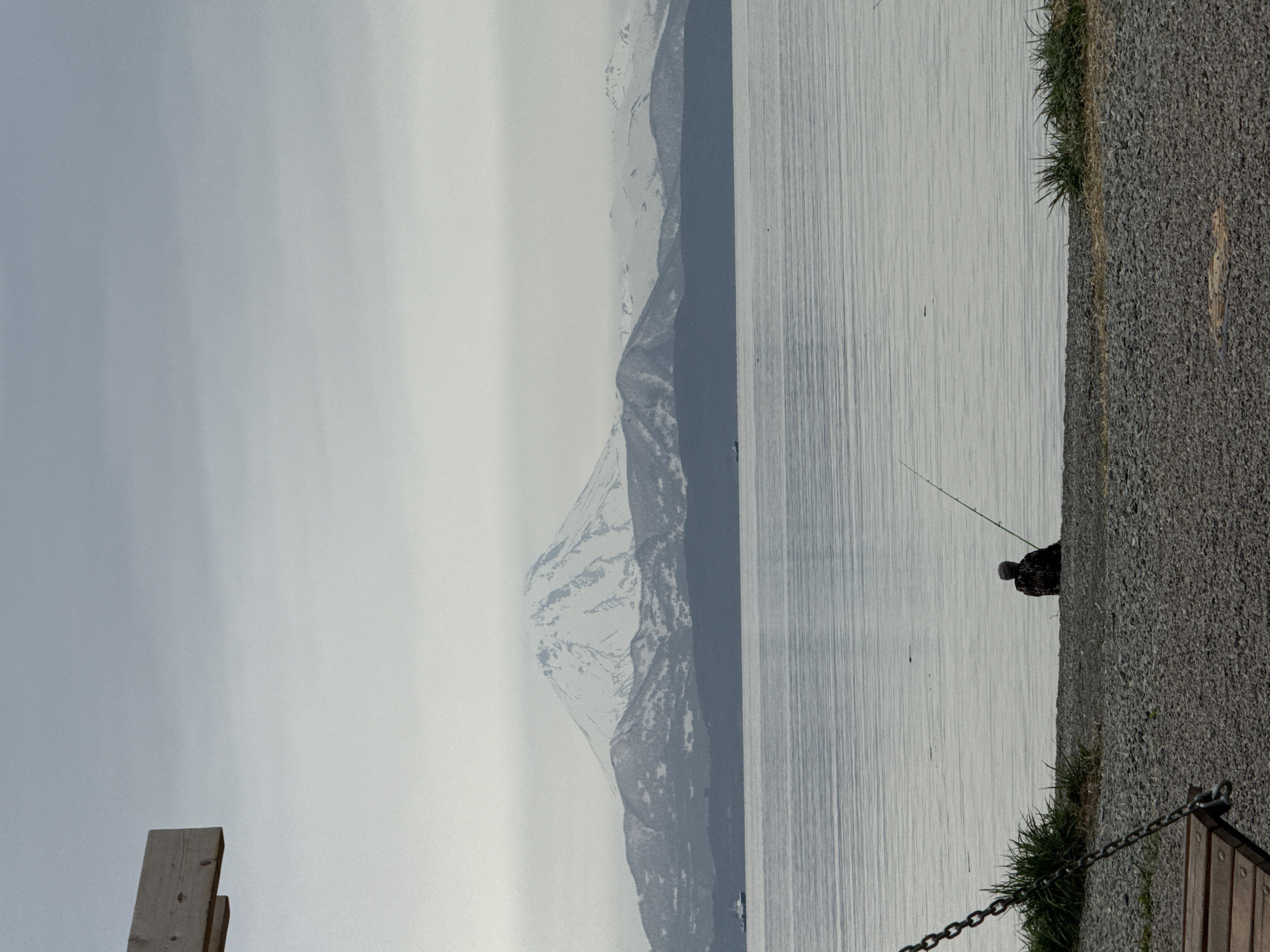 The image depicts a serene coastal scene in Petropavlovsk-Kamchatsky, Russia, with a snow-capped volcano looming in the background. In the foreground, a lone figure sits on a pebble-strewn shore, engrossed in fishing with a rod. The calm waters of the bay gently lap the shore, reflecting the overcast sky. The presence of a swing set with wooden planks and metal chains suggests a recreational area, possibly a park or a viewpoint overlooking the bay. The overall atmosphere is one of tranquility and solitude, offering a glimpse into the natural beauty of this remote Russian city.