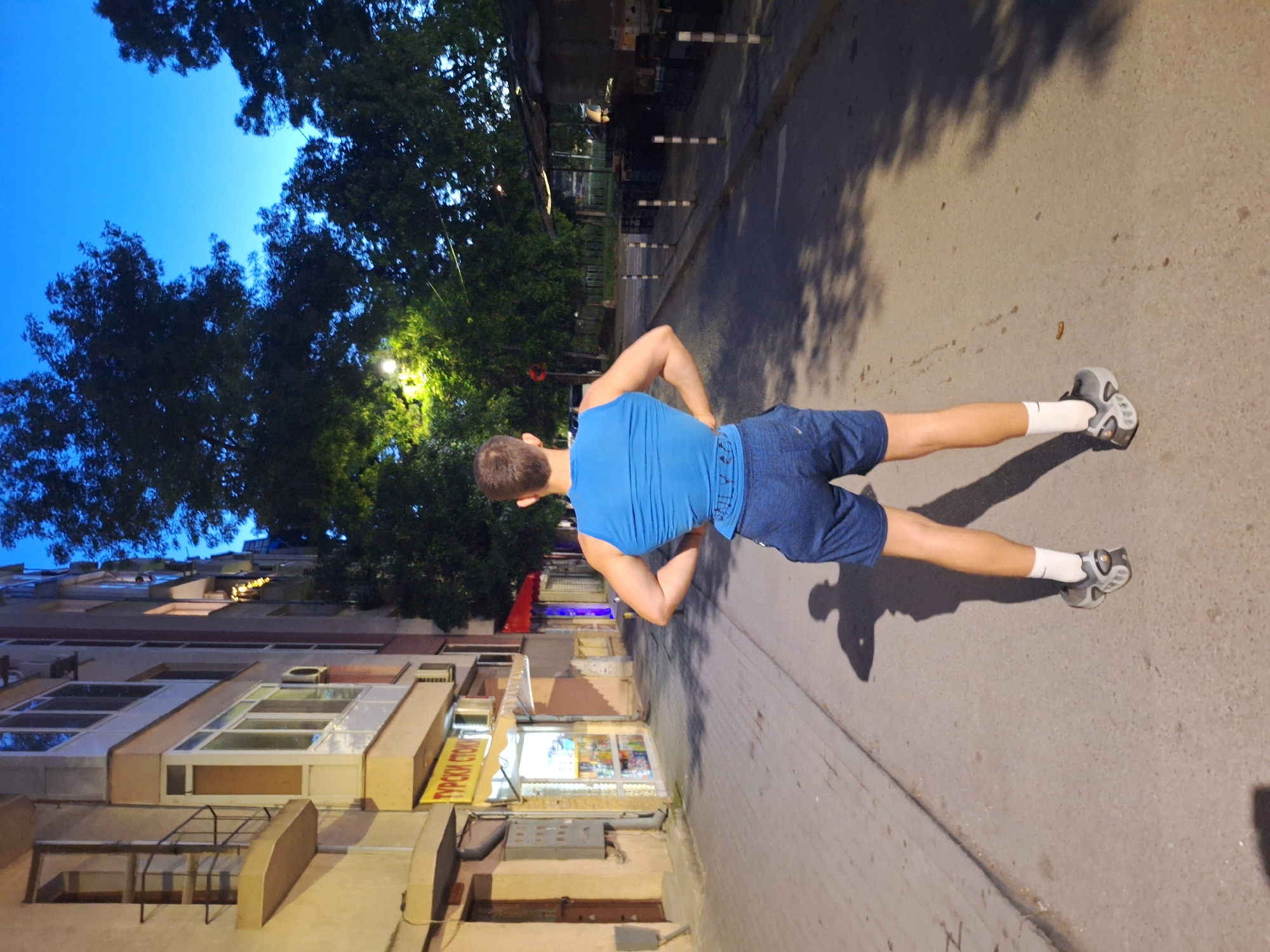 The image depicts a young man standing with his back to the camera, facing a street in Varna, Bulgaria. He is wearing a blue sleeveless shirt and dark blue shorts, with white socks and grey athletic shoes. The street is paved and appears to be a sidewalk or pedestrian area, lined with buildings on one side and trees and a makeshift market stall on the other. The buildings are of varying heights and architectural styles, with windows and balconies visible. One building has a sign in Cyrillic script reading "TYPCKИ CTOKH," indicating a shop selling Turkish goods. The sky is a deep blue, suggesting either early morning or late evening. The lighting is dim, with streetlights casting shadows on the ground, creating a somewhat dramatic atmosphere. The man's posture, with hands on his hips, suggests a moment of pause or contemplation. The overall scene conveys a sense of urban life in Varna, with a blend of residential and commercial elements, and a touch of the natural environment provided by the trees.
