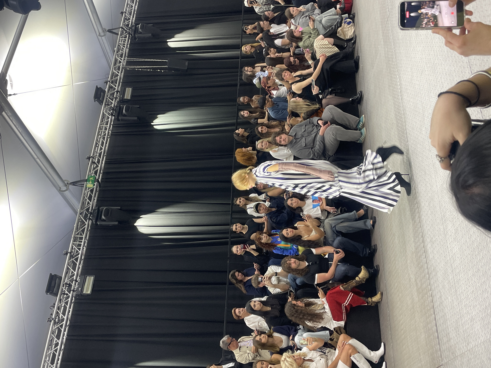 This image captures an indoor runway event, likely a fashion show, taking place in Barcelona, Spain. The scene features a light-colored, textured runway with rows of seated spectators lining both sides.
In the midground, a person, presumably a model, is walking down the runway. They are wearing a striking long dress with wide horizontal stripes in black, white, and a light blue or grey. The model has blonde hair.
The audience, consisting of numerous individuals of varying ages and styles, is closely seated, facing the runway. Many spectators are actively engaged, watching the model, and a significant number are holding up smartphones to record or photograph the event. In the immediate foreground, a pair of hands holding a smartphone is visible, indicating the photograph was taken from an attendee's perspective, capturing the show on their device. One audience member can be seen wearing a white t-shirt with a printed face.
The setting is an indoor space, possibly a large hall or tent, with a dark, pleated curtain serving as a backdrop on the left side of the runway. Above this backdrop, a metal truss structure is visible, equipped with spotlights and other lighting fixtures that illuminate the event. A green "Exit" sign with a white arrow pointing left and a person icon is affixed to the truss. The ceiling appears to be a light-colored, possibly fabric, structure with bright, artificial overhead lighting. The atmosphere suggests a lively and well-attended fashion presentation.
