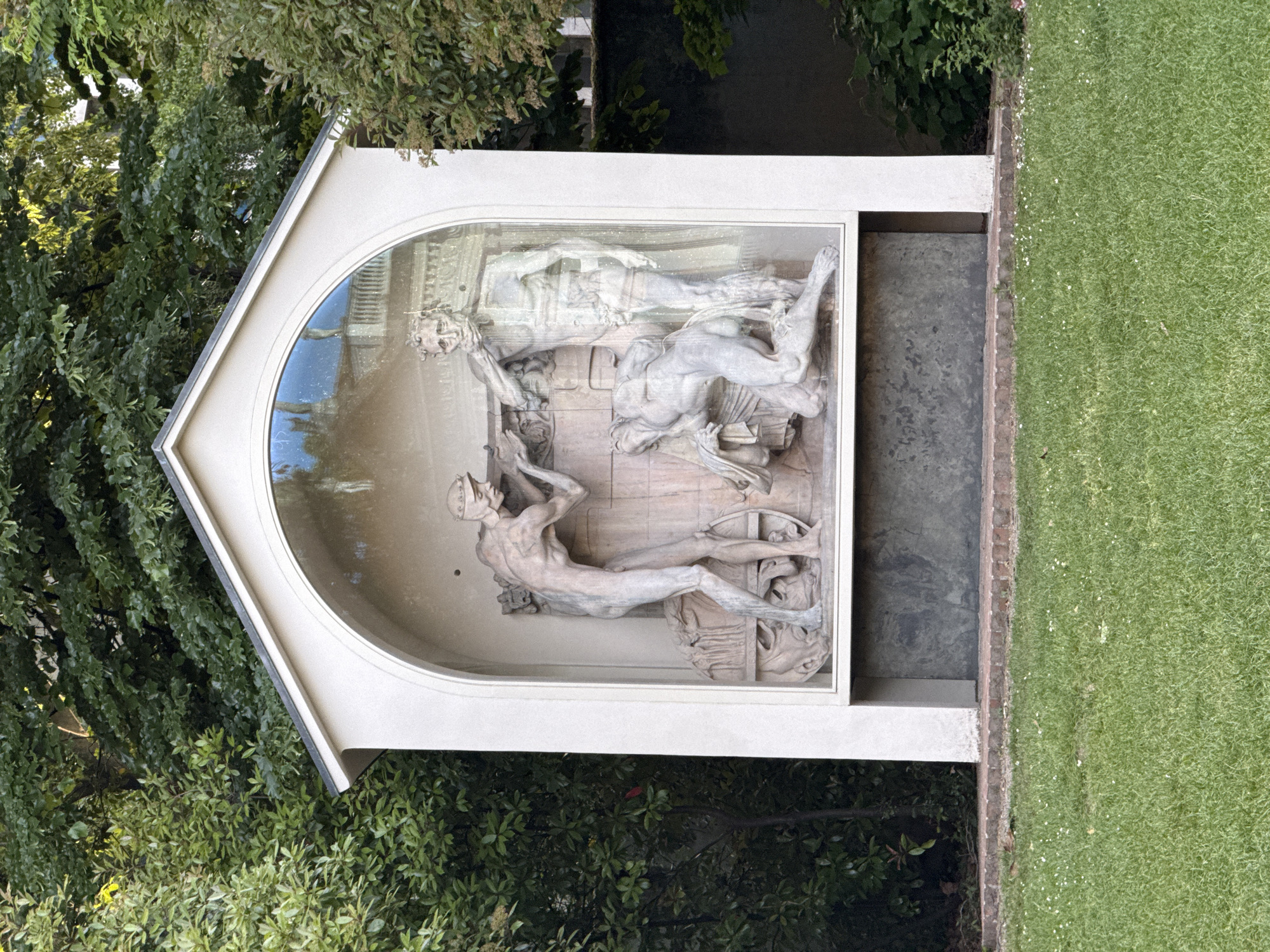 The image shows a monument in Milan, Italy. The monument is a white structure with a gable roof, and it houses a sculpture depicting several nude male figures in a classical style. The sculpture is housed behind glass and appears to be in a state of disrepair. The monument is surrounded by greenery, including trees, bushes, and a lawn. The monument is likely located in a park or garden setting.