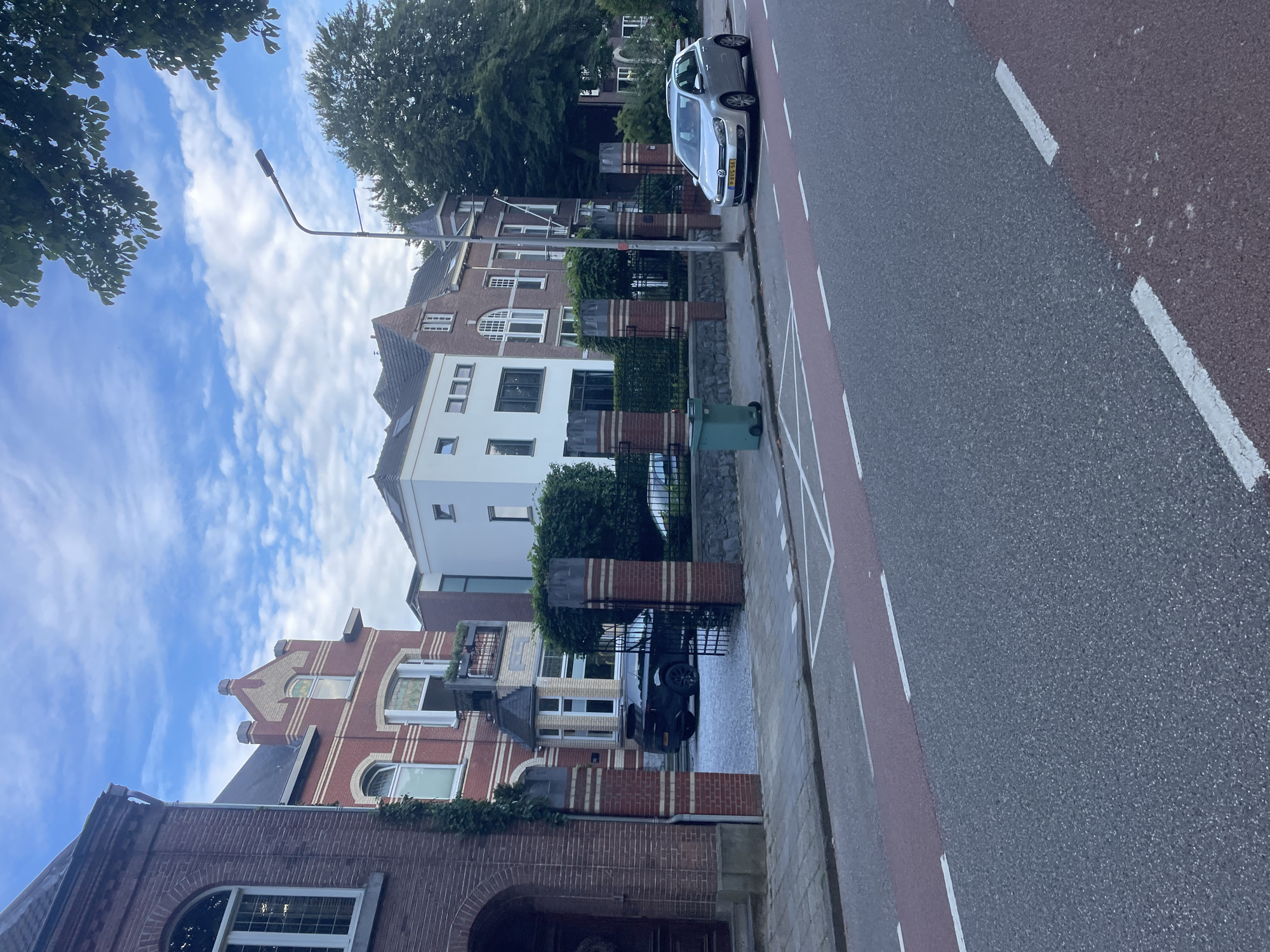 This media file shows a street scene in Weert, Netherlands. The perspective is from a low angle looking up at the sky and then down the street, with the camera rotated.
The subjects include several residential buildings of varying architectural styles, characterized by brickwork and white facades. There is a silver Volkswagen Golf parked on the right side of the street, facing away from the viewer. A black Mini Cooper is parked in the driveway of one of the houses. A green trash bin is visible on the sidewalk. Trees line the street and are visible in the background. The sky is mostly blue with scattered white clouds.
The scene appears to be during the daytime, likely late morning or early afternoon, given the lighting. The weather seems pleasant, with clear skies and daylight. The location is identifiable as a residential street in the Netherlands, indicated by the car's license plate, which is a Dutch format. There is no explicit activity or event taking place, suggesting a static street view. The overall impression is one of a quiet, everyday street in a European town.