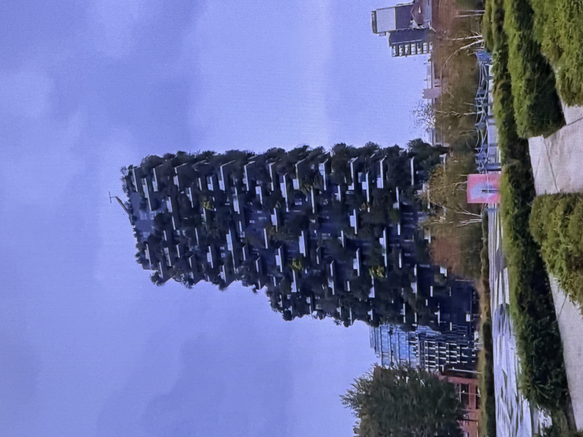 The image depicts a striking, modern high-rise building with a unique, tiered design covered in lush greenery, resembling a vertical forest. The building's facade is characterized by numerous balconies adorned with trees and plants, giving it a living, organic appearance that contrasts with its man-made structure.
To the right of the building, there is a pathway or sidewalk lined with neatly manicured hedges, suggesting a well-maintained urban park or public space. Further in the background, other buildings are visible, indicating an urban environment. One of these buildings appears to be a modern skyscraper with reflective glass windows. A pink banner with a white oval shape is also visible in the mid-ground, adding a splash of color to the scene.
The sky is overcast with a muted, pale blue hue, suggesting it might be a cloudy day or perhaps early morning or late afternoon. The overall lighting is soft and diffused.
The scene appears to be an outdoor setting, possibly a city square or a park adjacent to the residential or commercial building. There are no people visible in the image, and the focus is entirely on the architecture and landscape. The prominent building with its integrated vegetation is the central subject, showcasing an innovative approach to urban design and sustainable living.
The location is identified as Dubai, United Arab Emirates, which adds context to the architectural style and the presence of such modern, ambitious structures in a desert climate. The building's design, with its extensive planting, is particularly noteworthy given Dubai's environment, highlighting efforts towards green spaces and biodiversity in the city.