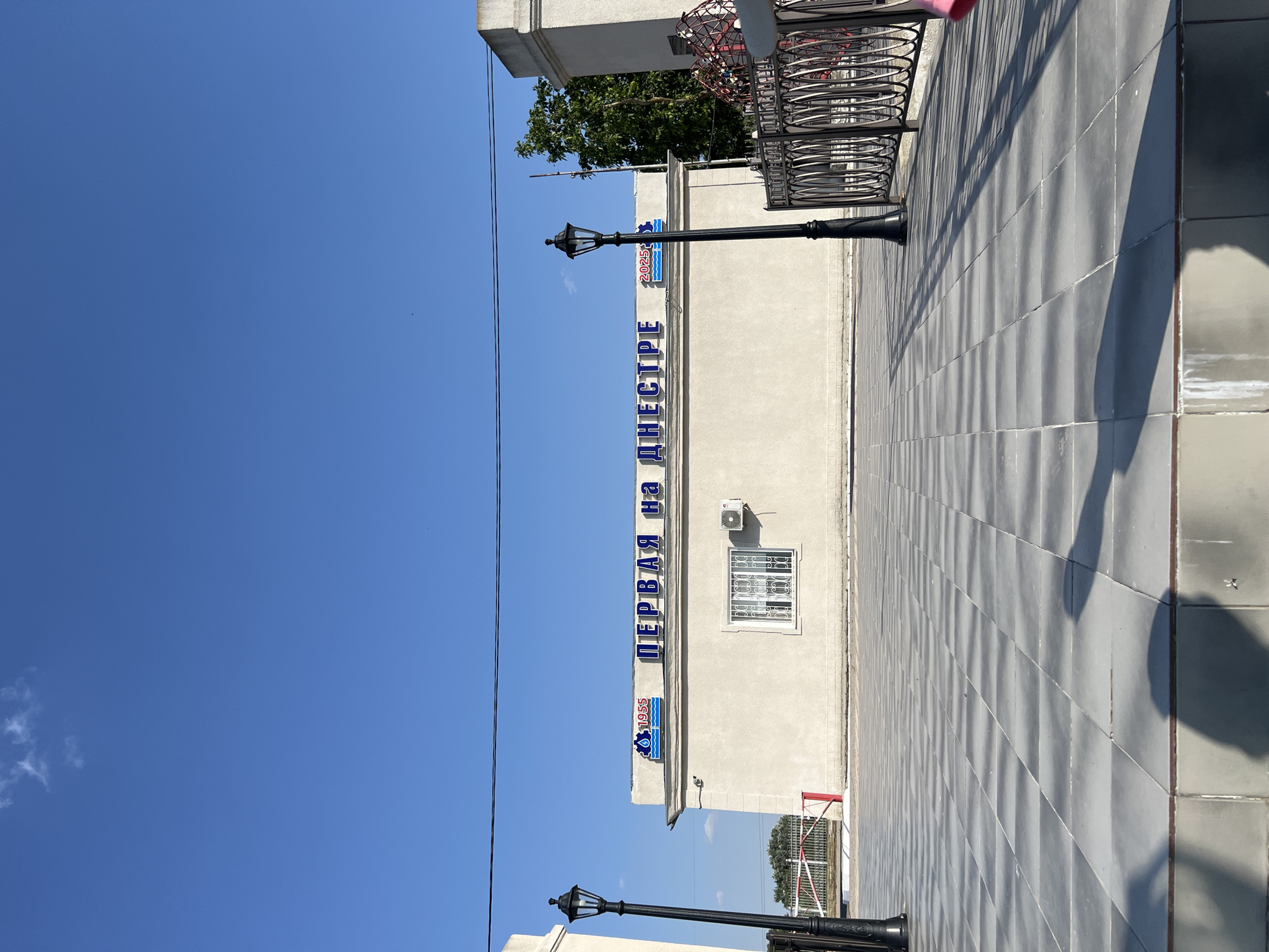 A low-angle, full shot captures a bright, sunny day with a clear blue sky in Korzhevo, Moldova. In the center of the frame, a one-story, light-colored building is visible. Blue Cyrillic lettering that reads "ПЕРВАЯ НА ДНЕСТРЕ" (First on the Dniester) is prominently displayed across the top of the building's facade. To the left of the lettering, there is a logo with the number "1955" and a graphic of water. To the right, there is another logo with the number "2025" and a graphic of water. A white-framed window with ornate metal bars is centered on the building's facade, and an air conditioning unit is mounted above it. To the left and right of the building, two black lampposts with ornate designs stand tall. The ground in front of the building is paved with large, gray square tiles arranged in a checkerboard pattern, casting long shadows. In the foreground, shadows of two people are cast on the tiled ground, suggesting their presence just outside the frame. A black metal railing with circular designs runs along the right side of the tiled area, partially obscuring a red structure in the background. A white gate with red and white striped poles is visible to the left of the building, behind a short fence. Trees can be seen behind the fence line. The overall scene appears to be an outdoor public space, possibly a plaza or walkway, leading up to the building.
