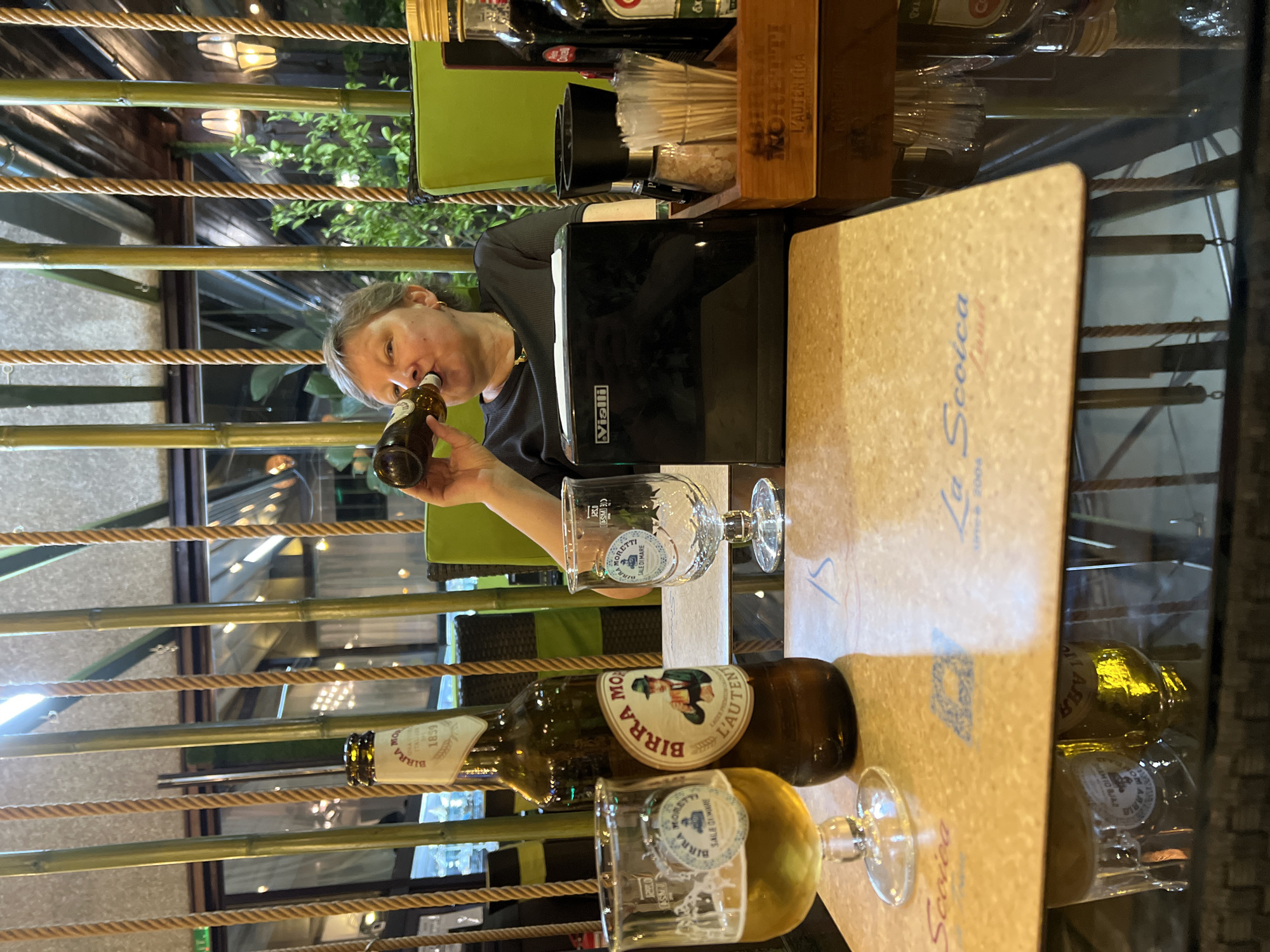 The image shows a woman drinking from a beer bottle at a table in what appears to be an outdoor seating area of a restaurant. The table has a coaster with the text "La Scoica" and what looks like "since 2004," suggesting the location is La Scoica in Constanța, Romania. On the table are two beer glasses, one with beer, a bottle of Birra Moretti, a salt and pepper grinder, and a box containing toothpicks. Behind the table, bamboo stalks and rope decor are visible, and in the background, there are lights and greenery, indicating an evening setting. The woman is wearing a dark top and a necklace, and her short grey hair is visible.